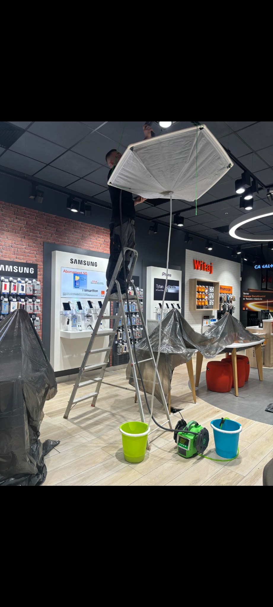 Pracownik na drabinie montuje lub naprawia instalację podwieszaną pod sufitem w salonie sprzedaży, zabezpieczone folią meble, zielony i niebieski pojemnik oraz urządzenie do czyszczenia.