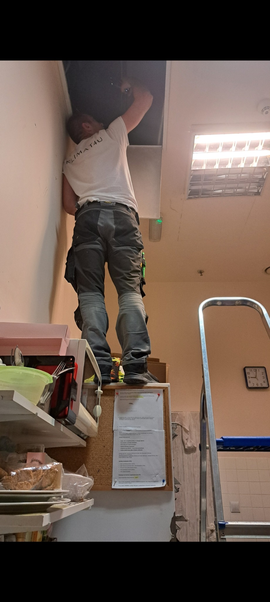 Pracownik w firmowej koszulce KLIMATA4U stoi na szafce, naprawiając lub instalując coś w suficie podwieszanym, widoczne narzędzia i drabina aluminiowa w tle.