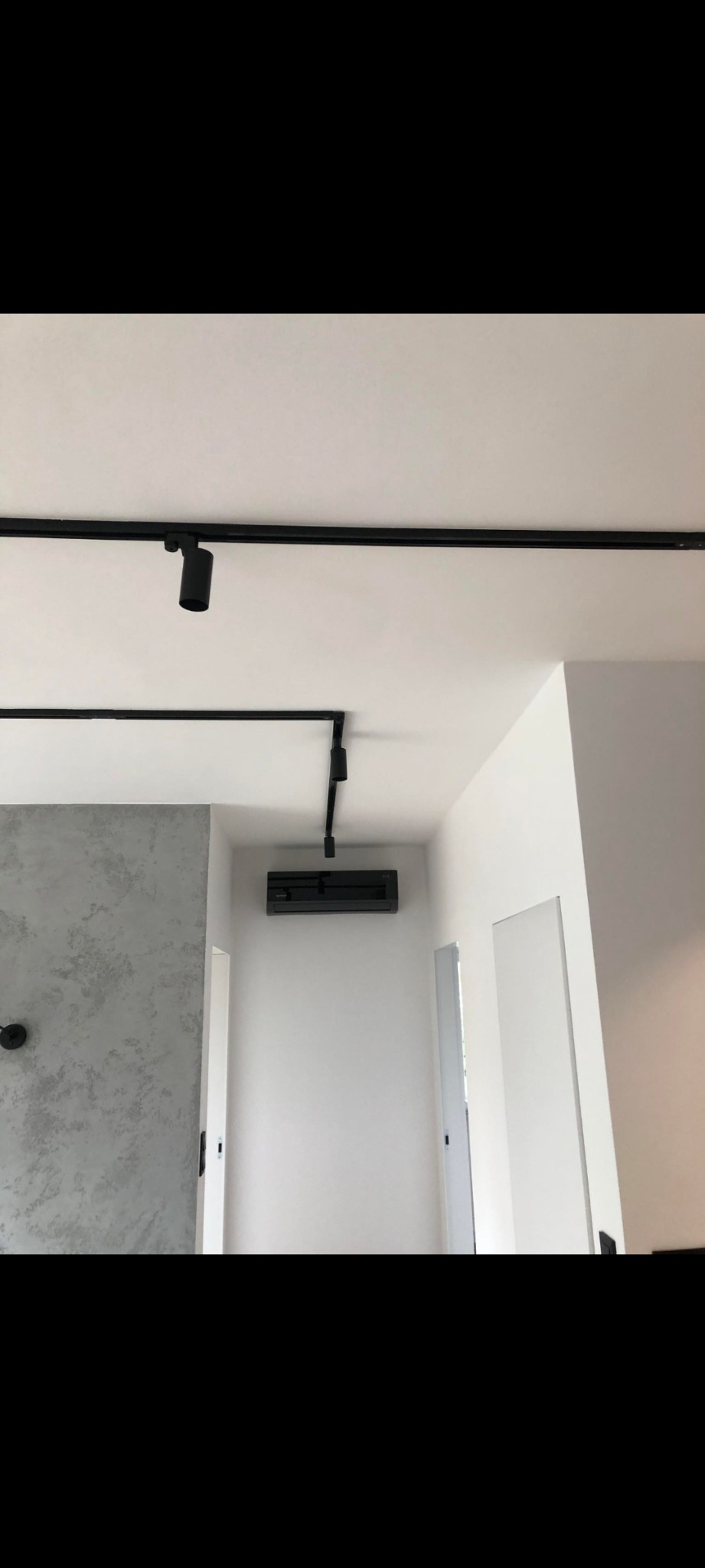 Nowoczesne wnętrze z szarą ścianą o fakturze betonu, czarnym oświetleniem szynowym i zamontowaną klimatyzacją na białej ścianie.