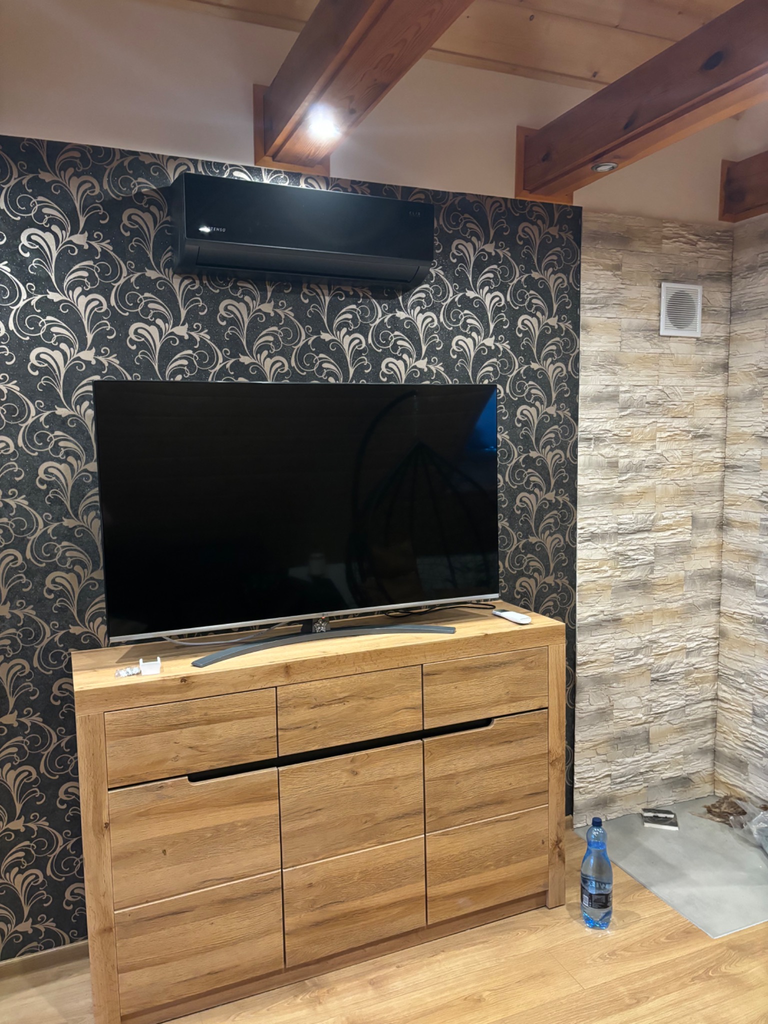 Czarny klimatyzator ścienny zamontowany nad drewnianą komodą pod telewizorem na tle ściany z wzorzystą tapetą, obok ściana wyłożona kamieniem dekoracyjnym, widoczne drewniane belki stropowe.