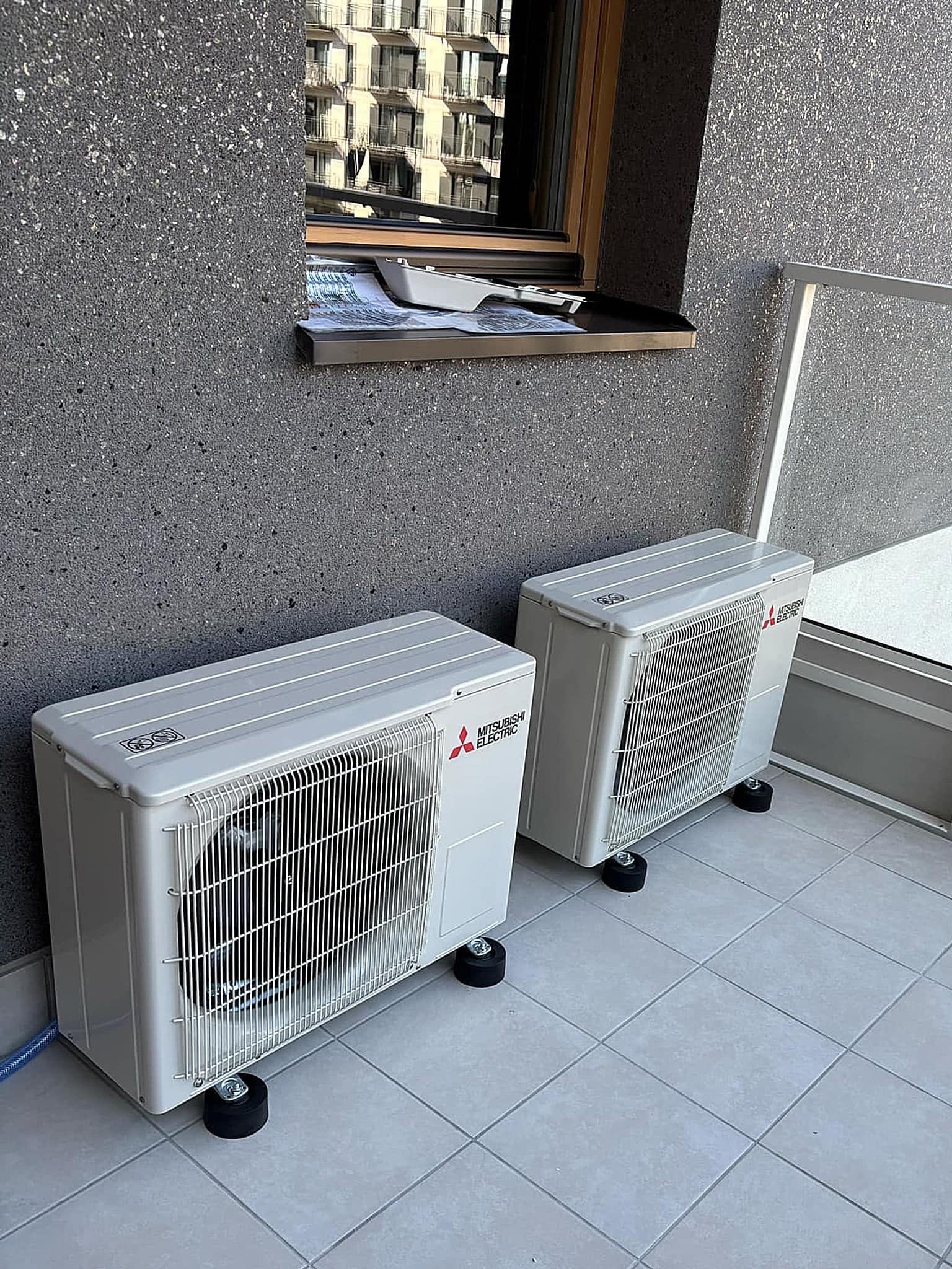 Dwa białe agregaty klimatyzacji Mitsubishi Electric ustawione na czarnych podkładkach antywibracyjnych na balkonie wyłożonym szarą terakotą, widok na budynek mieszkalny w tle.