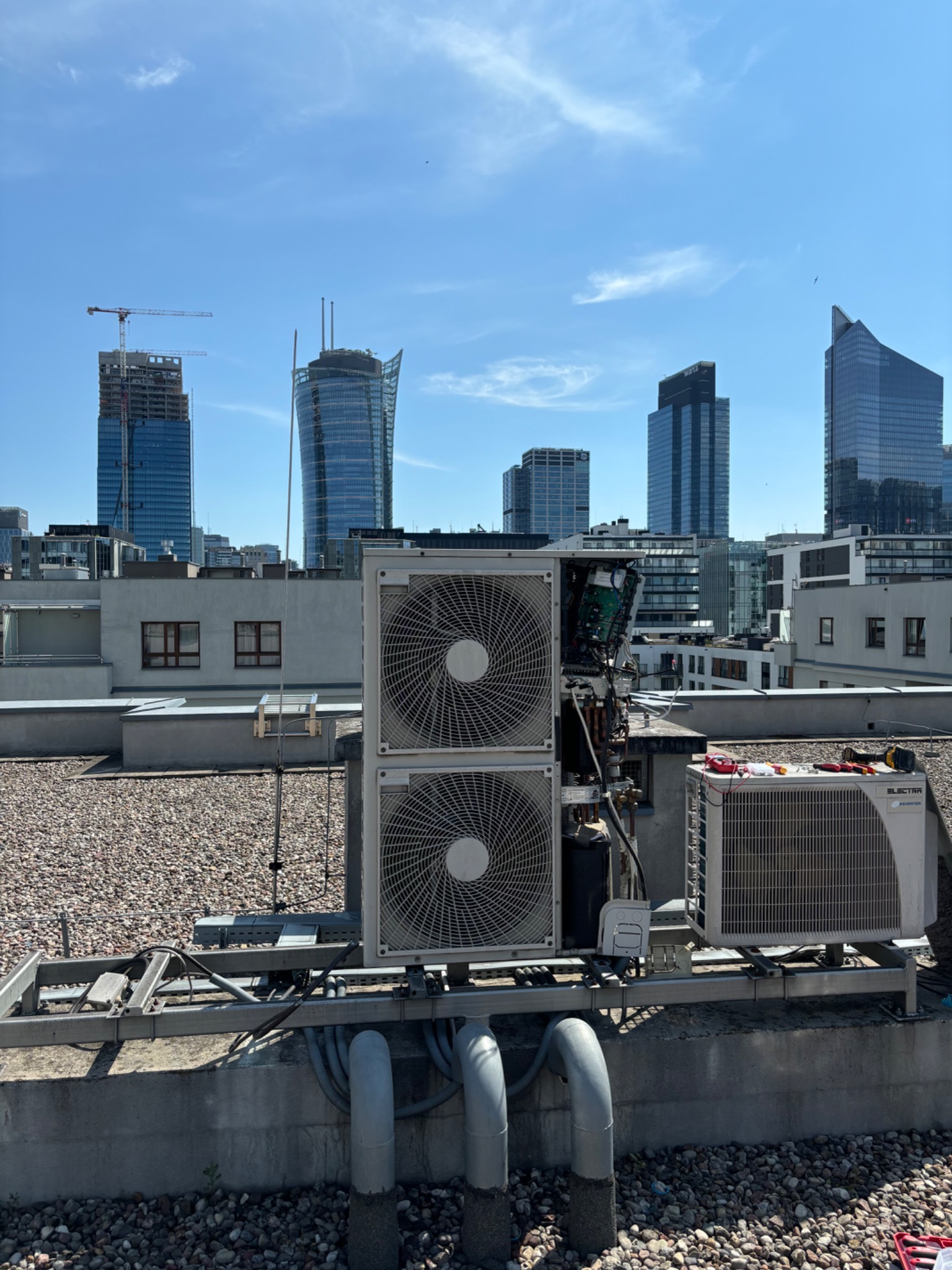 Dwie jednostki klimatyzacyjne na dachu budynku z odsłoniętą elektroniką jednej z nich, narzędzia obok, widok na panoramę miasta z drapaczami chmur w tle.