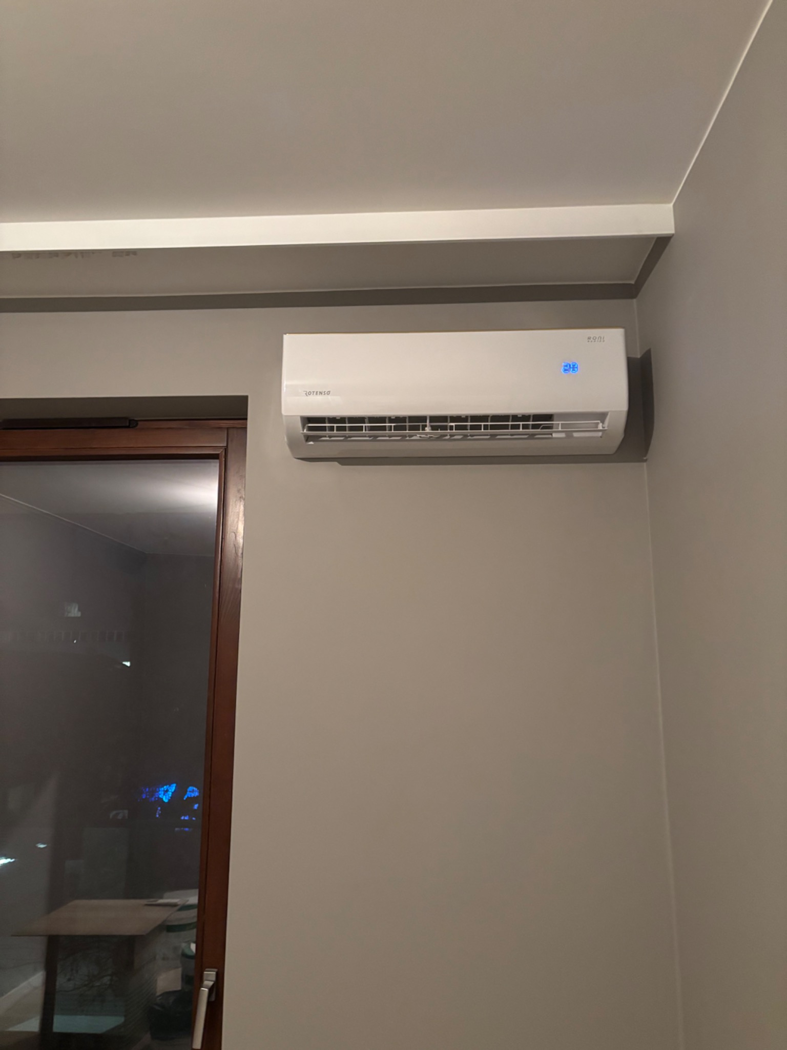 Biały klimatyzator ścienny marki Rotenso z wyświetlaczem temperatury, zamontowany w pokoju z szarą ścianą i brązową ramą okienną.