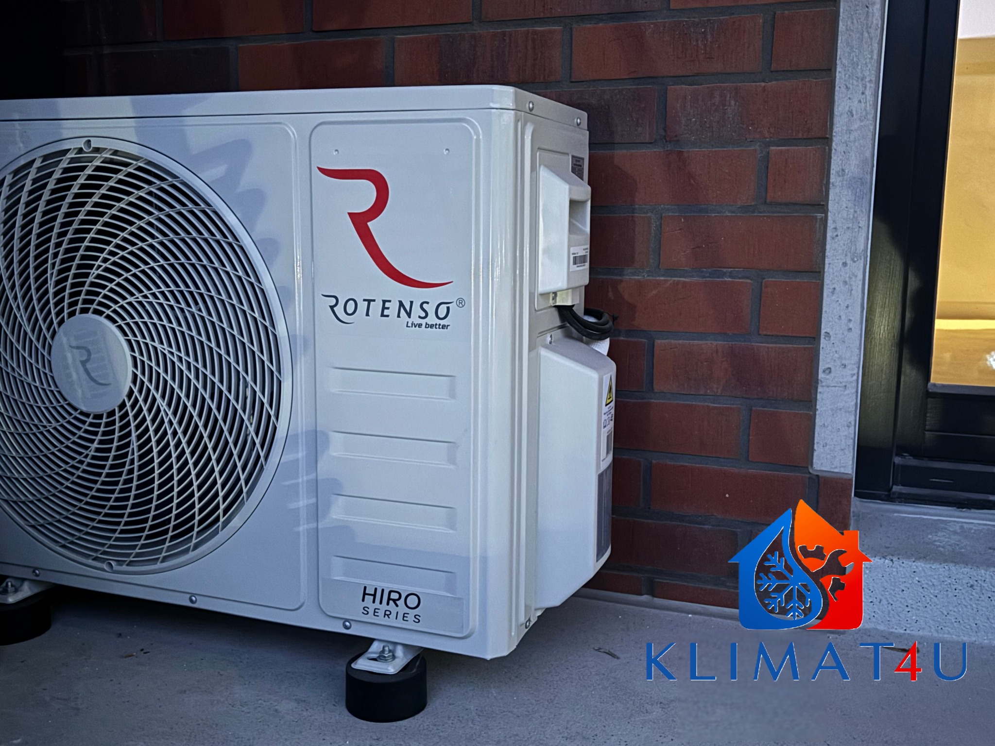 Zewnętrzna jednostka klimatyzacji Rotenso Hiro Series zamontowana na gumowych nóżkach, przy ścianie z czerwonej cegły, z widocznym logo firmy Klimat4U.