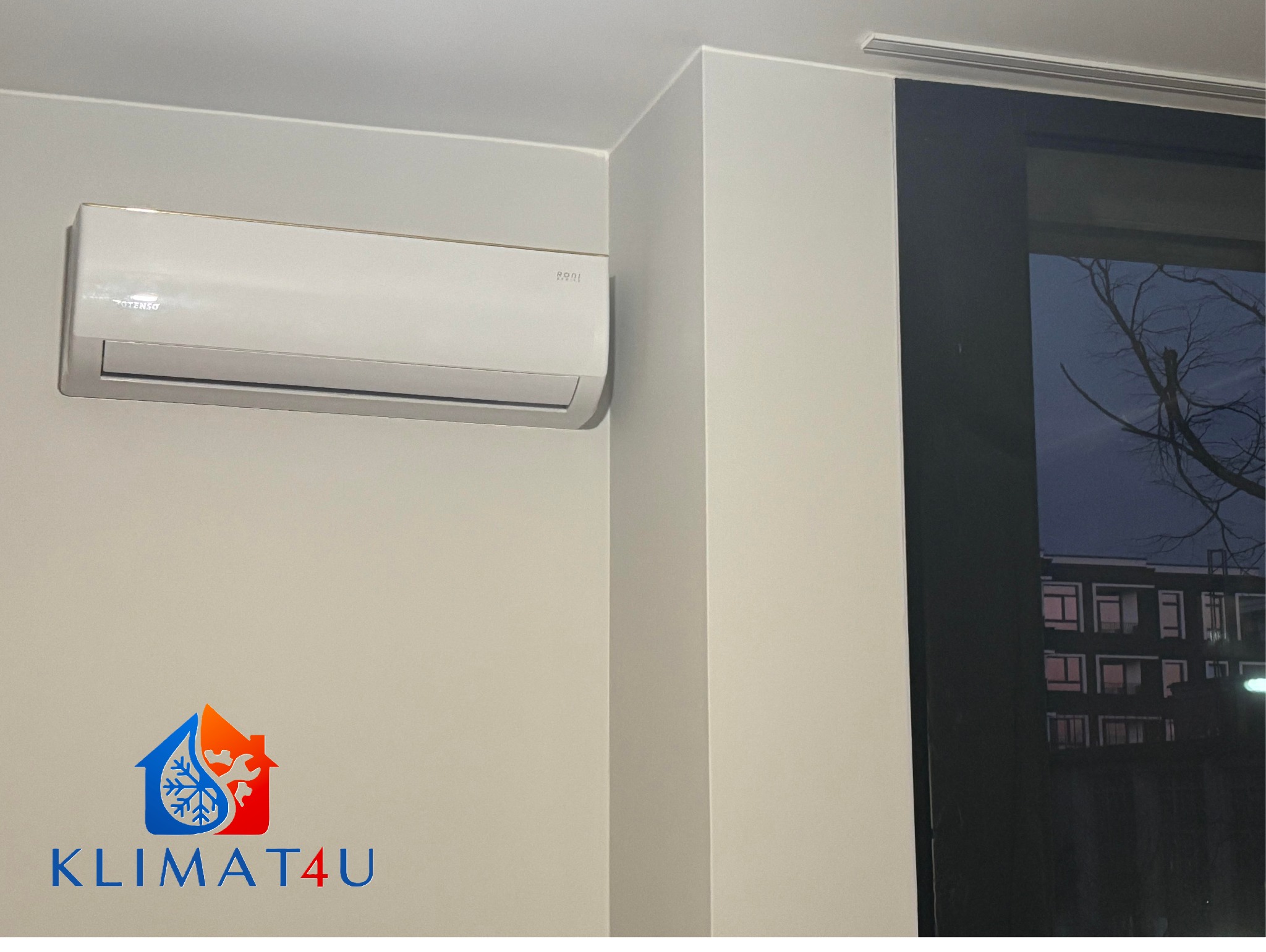 Biały klimatyzator z widocznym logo firmy na ścianie w pomieszczeniu z oknem, za którym widać budynek mieszkalny i nagie gałęzie drzewa.