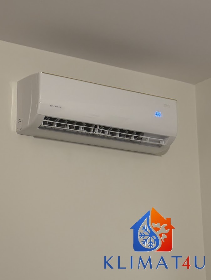 Biały klimatyzator ścienny z wyświetlaczem temperatury, zamontowany na jasnej ścianie w rogu pokoju, widoczne logo firmy na dole.