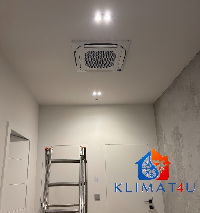 Kasetonowa jednostka klimatyzacji zamontowana w suficie podwieszanym, widoczna z dołu, z drabinką rozstawną opartą o ścianę i logo firmy w prawym dolnym rogu.