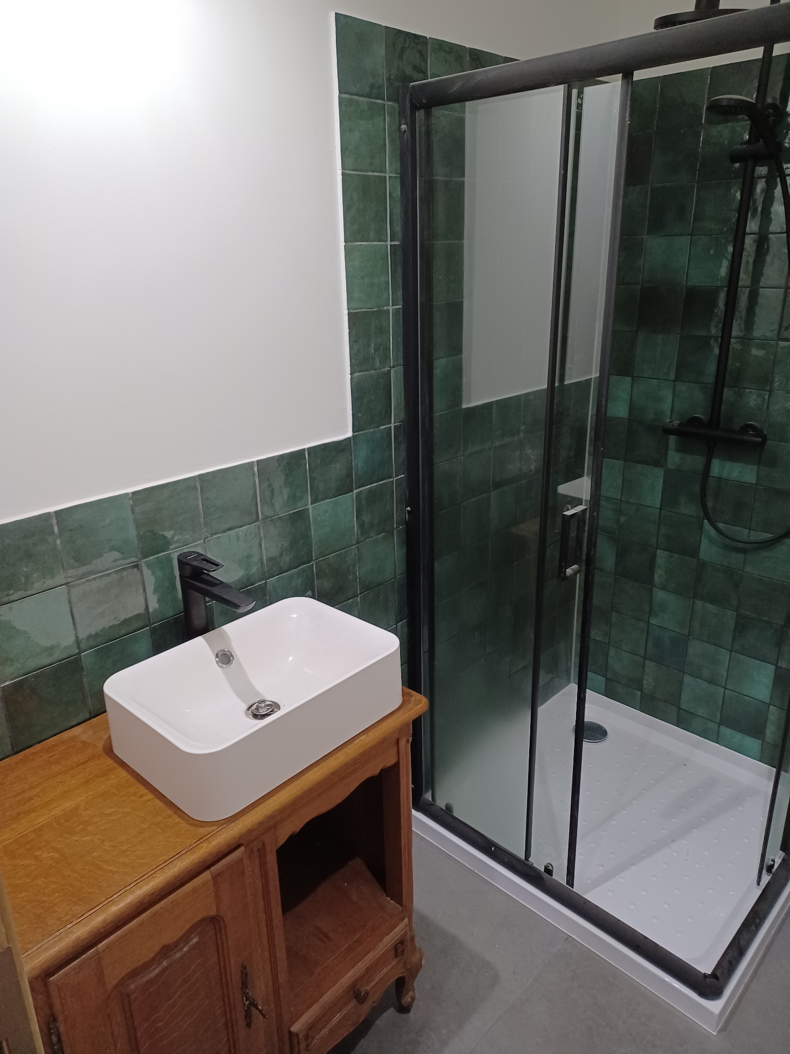 Nowoczesna łazienka z kwadratowymi, zielonymi płytkami, kabiną prysznicową z czarną ramą i umywalką nablatową na drewnianej szafce.