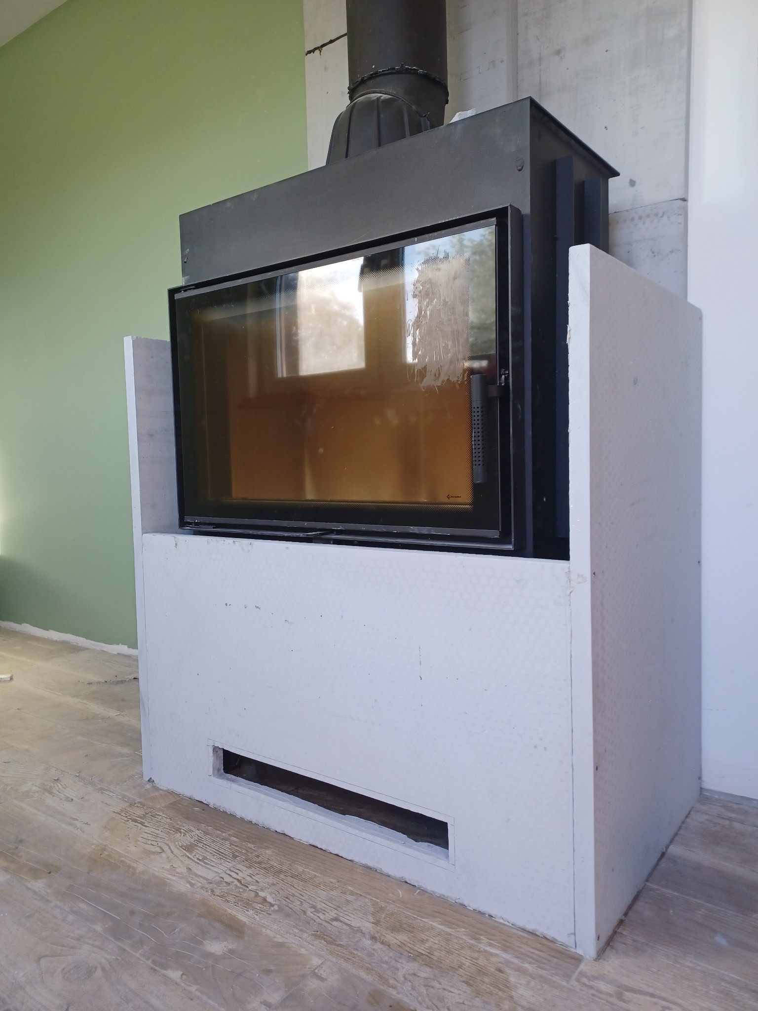 Wkład kominkowy z czarną ramą, obudowany surową, białą płytą gipsową z otworem wentylacyjnym u dołu, na tle zielonej ściany i surowego betonu, podłoga drewnopodobna.