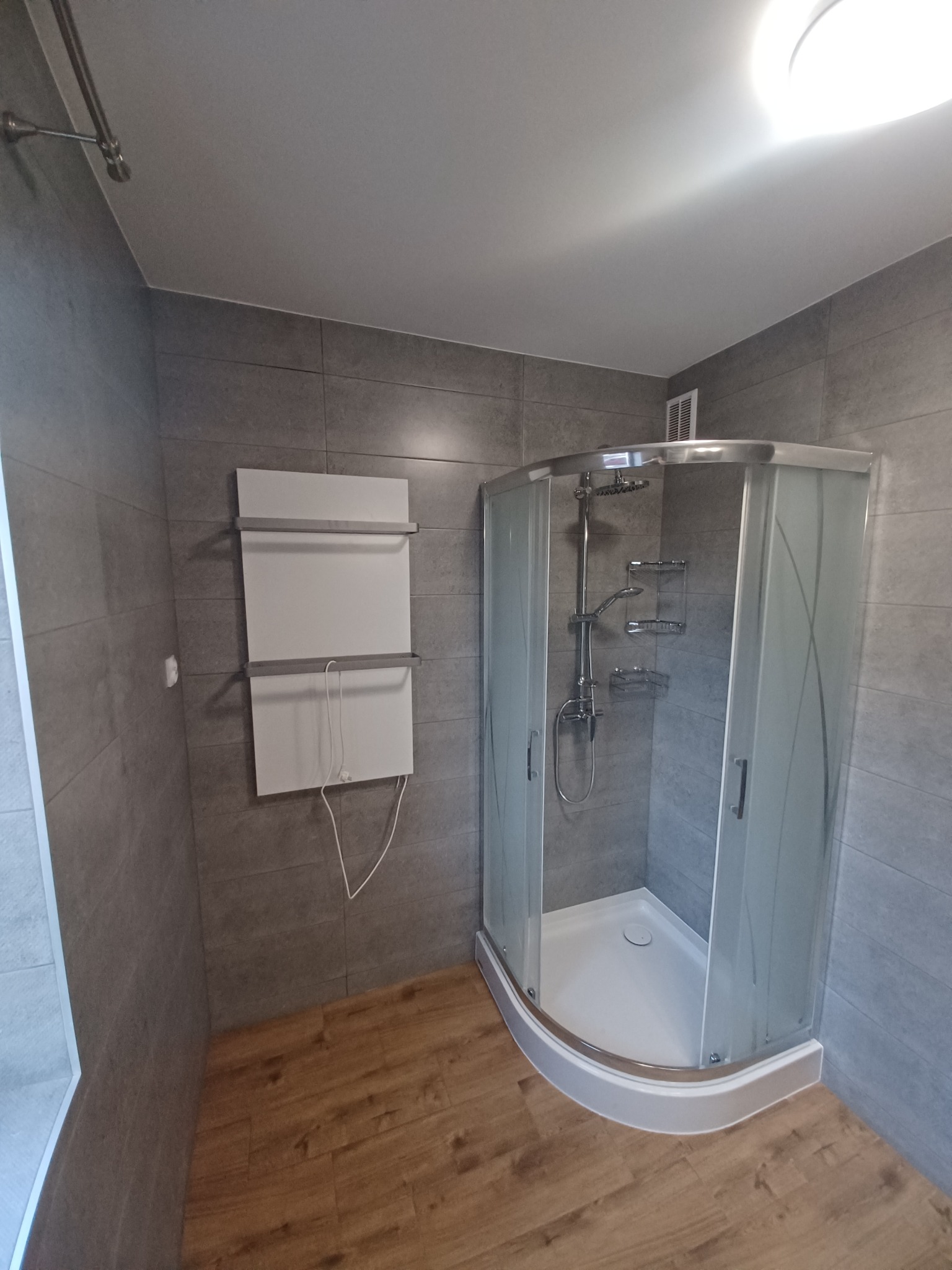 Nowoczesna kabina prysznicowa narożna z hydromasażem, szare płytki imitujące kamień na ścianach, drewnopodobna podłoga, grzejnik drabinkowy elektryczny na ścianie.