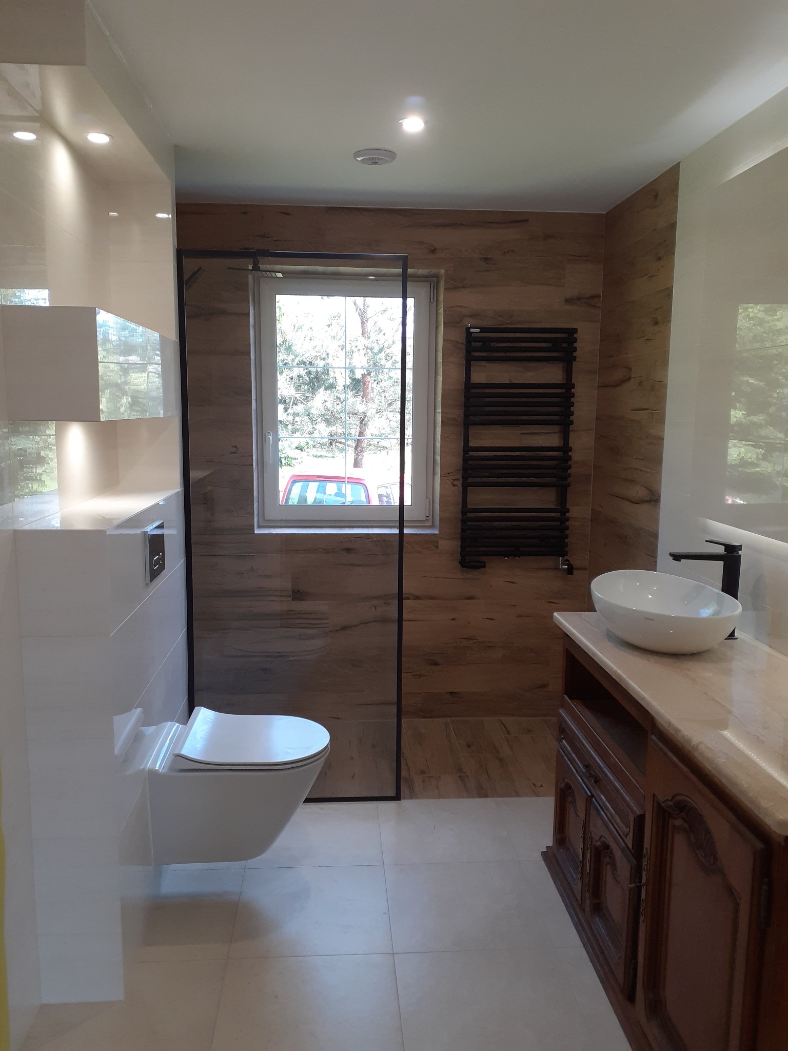 Nowoczesna łazienka z drewnopodobnymi płytkami na ścianie, wiszącą miską WC i grzejnikiem drabinkowym, oświetlona światłem dziennym wpadającym przez okno z widokiem na drzewa.