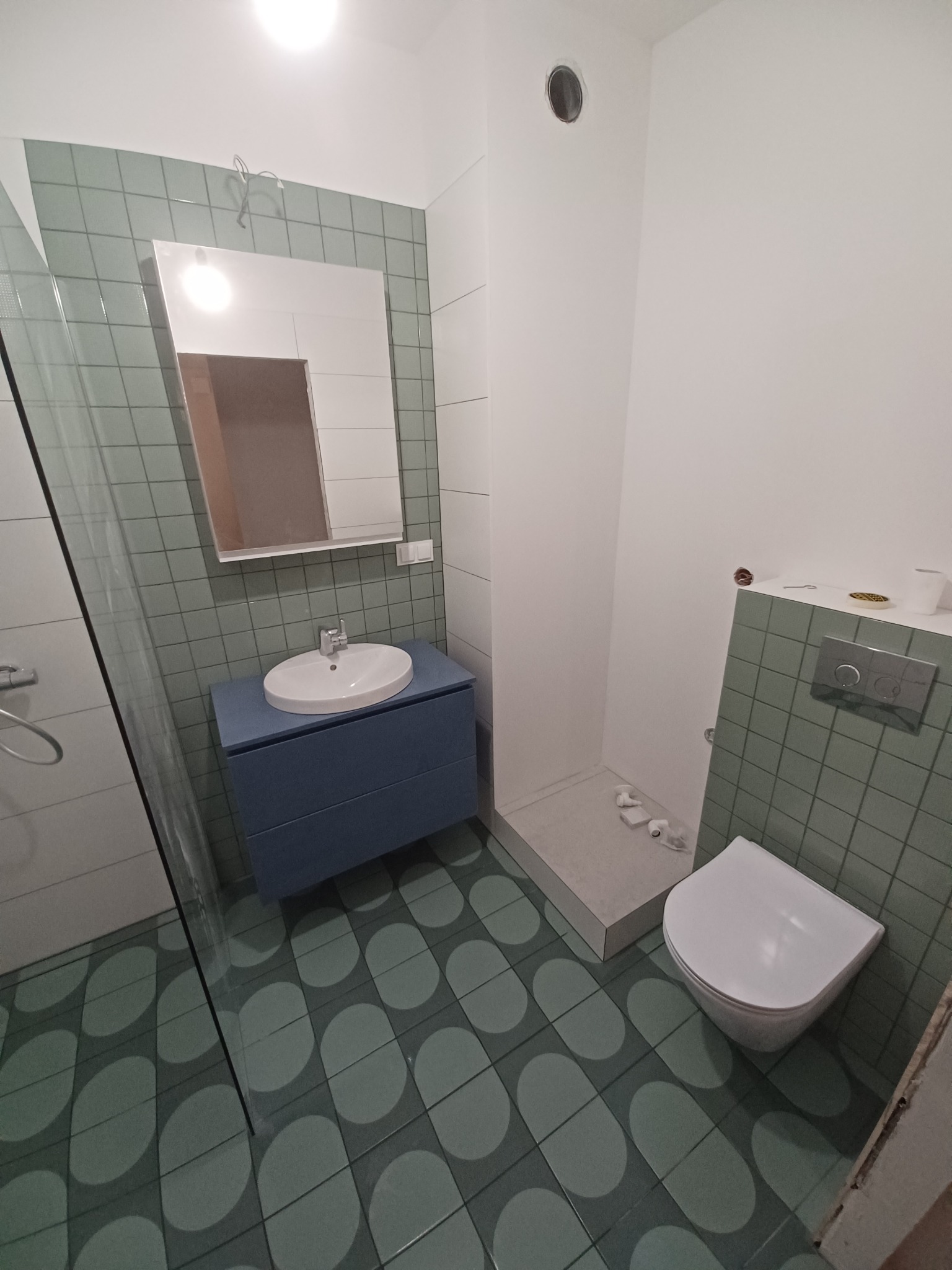 Wykończona łazienka w odcieniach zieleni z niebieską szafką pod umywalką, widoczne płytki podłogowe z motywem soczewek, biała toaleta i fragment kabiny prysznicowej.