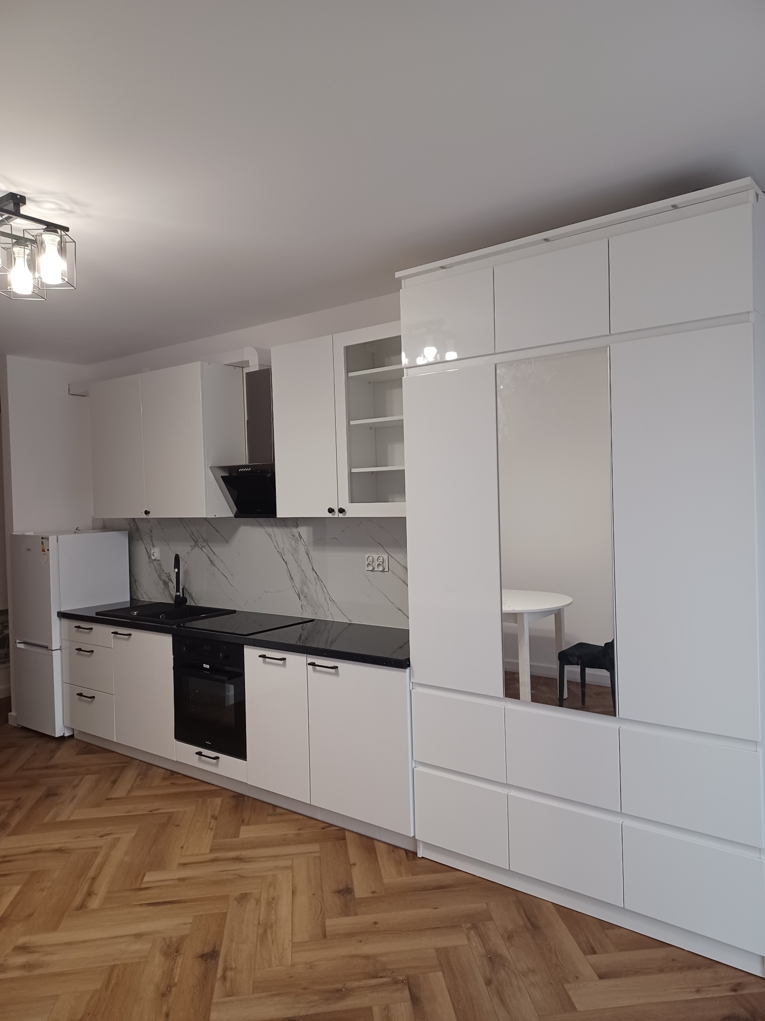 Nowoczesna, biała kuchnia z czarnym blatem i marmurowym panelem nad blatem, parkiet w jodełkę na podłodze, odbicie stołu i krzesła w lustrzanym froncie szafy.