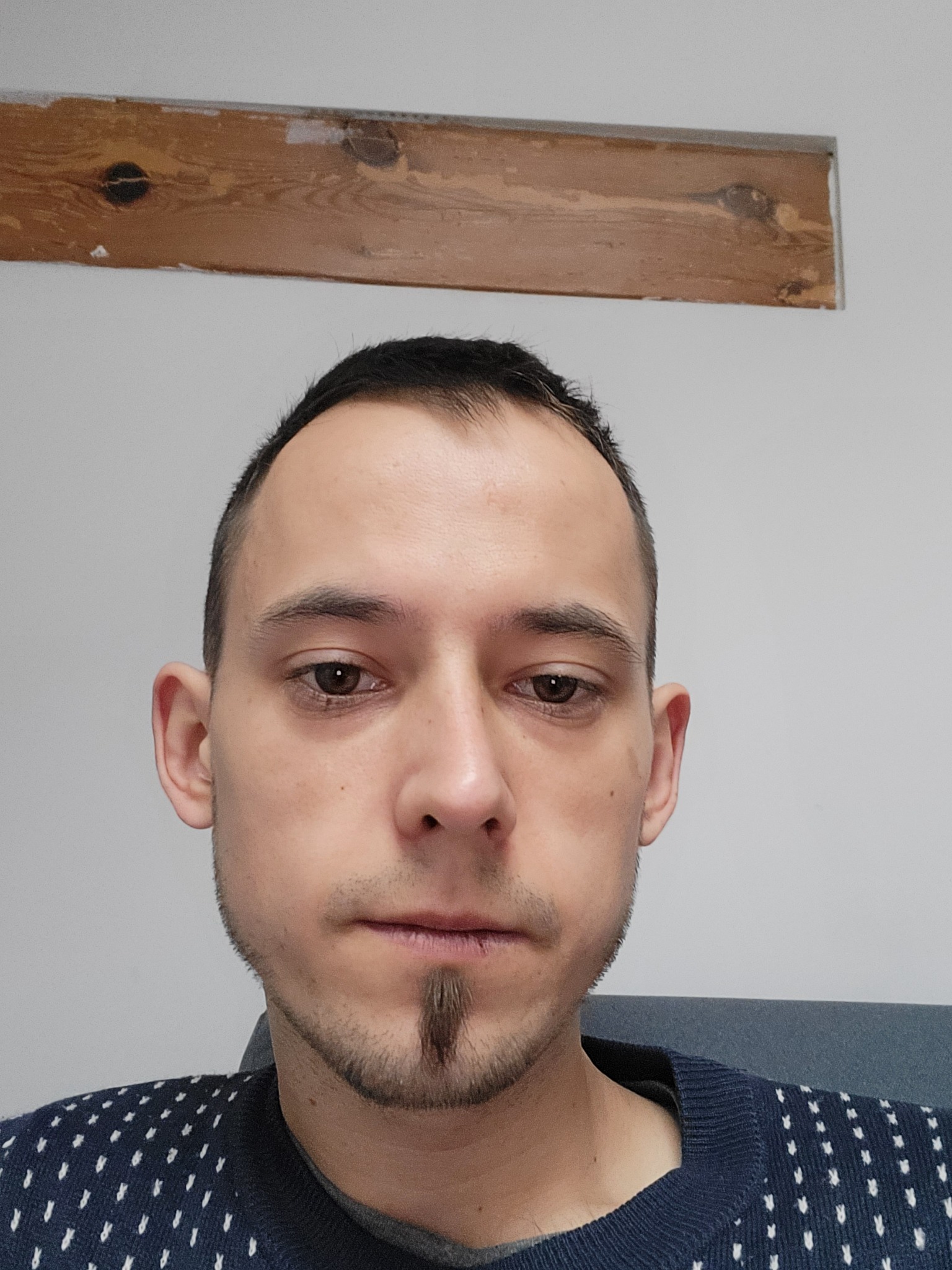 Portret mężczyzny z krótkimi włosami i kozia bródką, ubrany w granatowy sweter w białe wzory, na tle białej ściany z drewnianą listwą.