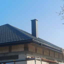 LEW-BUD - Niedokończony dom z ciemną dachówką i kominem. Widoczne rusztowania i drabina. Ściany z pustaków, częściowo otynkowane. Błękitne niebo w tle.