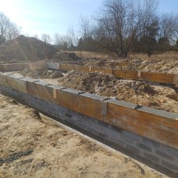 LEW-BUD - Wykopane fundamenty z drewnianym szalunkiem wypełnionym żwirem, widoczne bloczki fundamentowe i wystające pręty zbrojeniowe. W tle hałda ziemi i drzewa.