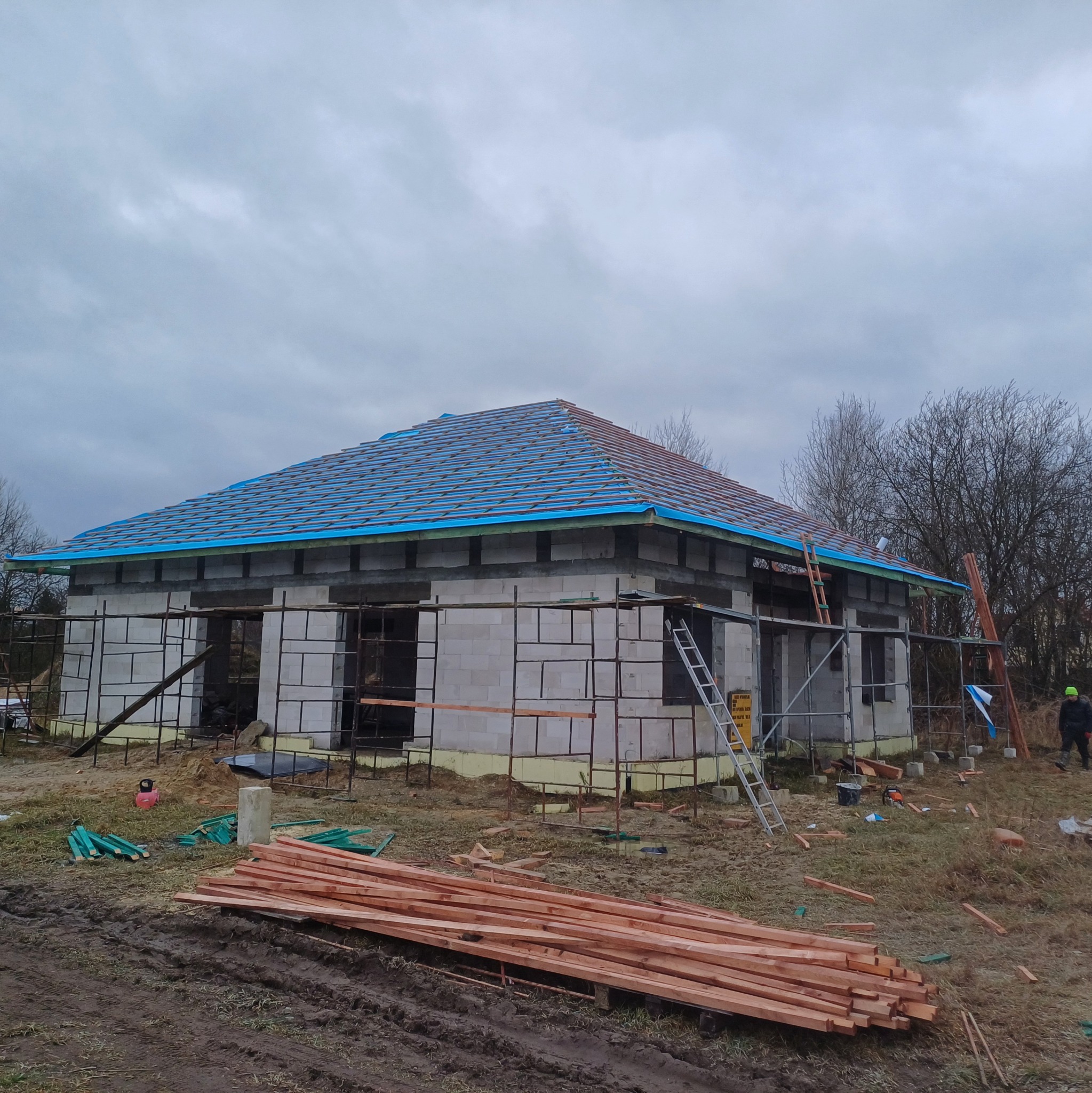 Budowa domu jednorodzinnego w stanie surowym otwartym. Widoczne ściany z bloczków, więźba dachowa z niebieską membraną i drewniane elementy konstrukcyjne.