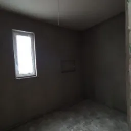 Surowe wnętrze pokoju po tynkowaniu maszynowym, widoczne okno z folią ochronną, ściany i sufit pokryte szarym tynkiem, mała wnęka w ścianie z wystającymi kablami.