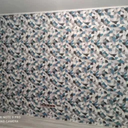 Ściana pokryta tapetą w geometryczny wzór z dominacją szarości, błękitu i czerni, z widocznymi gniazdkami elektrycznymi w dolnej części, oraz fragmentem drzwi po lewej stronie.