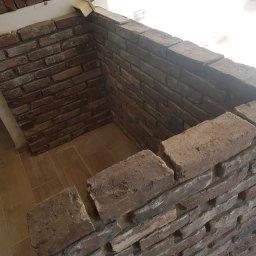 Michał Wojniusz - Fragment ściany wykończonej cegłą klinkierową w odcieniach brązu i szarości, z widoczną szczotką do fugowania na górnej krawędzi, na podłodze płytki imitujące drewno.