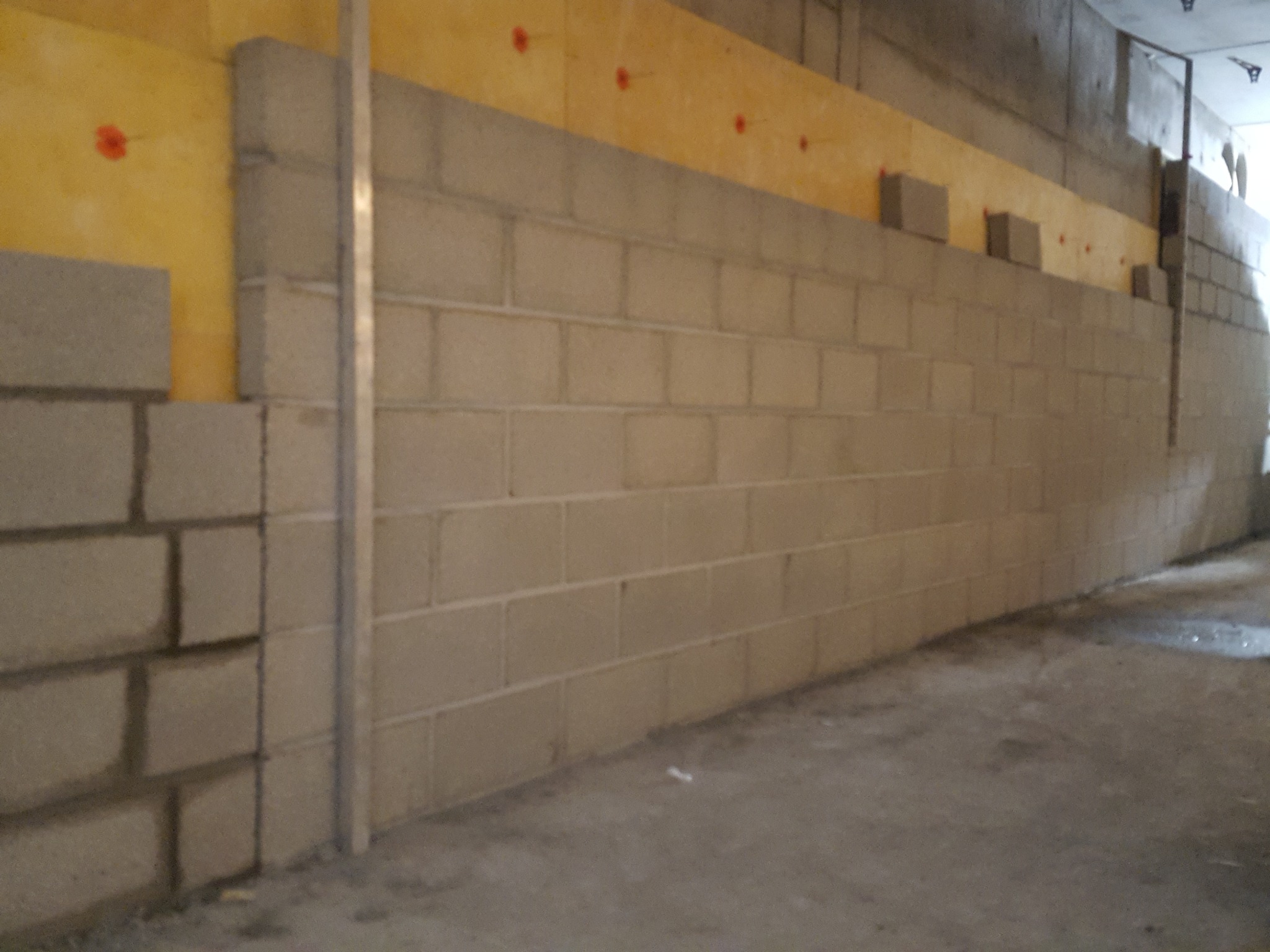 Wznoszenie ściany z bloczków betonowych na żółtej izolacji z widocznymi czerwonymi punktami mocowania.