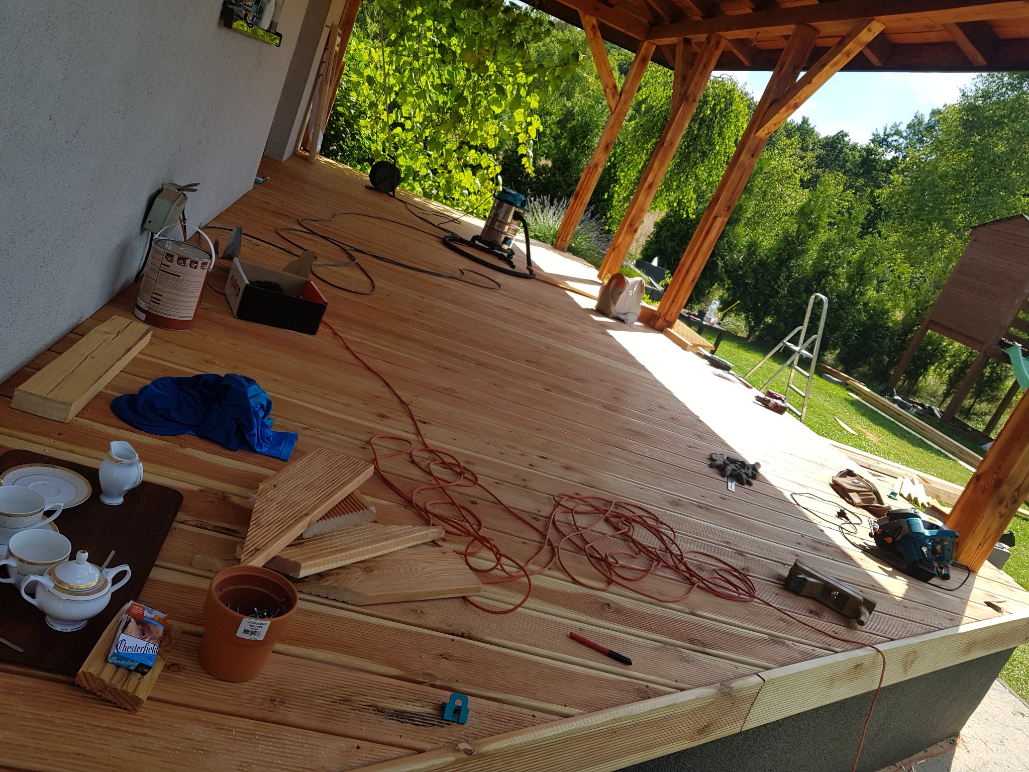 Montaż drewnianego tarasu: widok na deski, narzędzia, odkurzacz przemysłowy, oraz zestaw do herbaty na krawędzi tarasu.