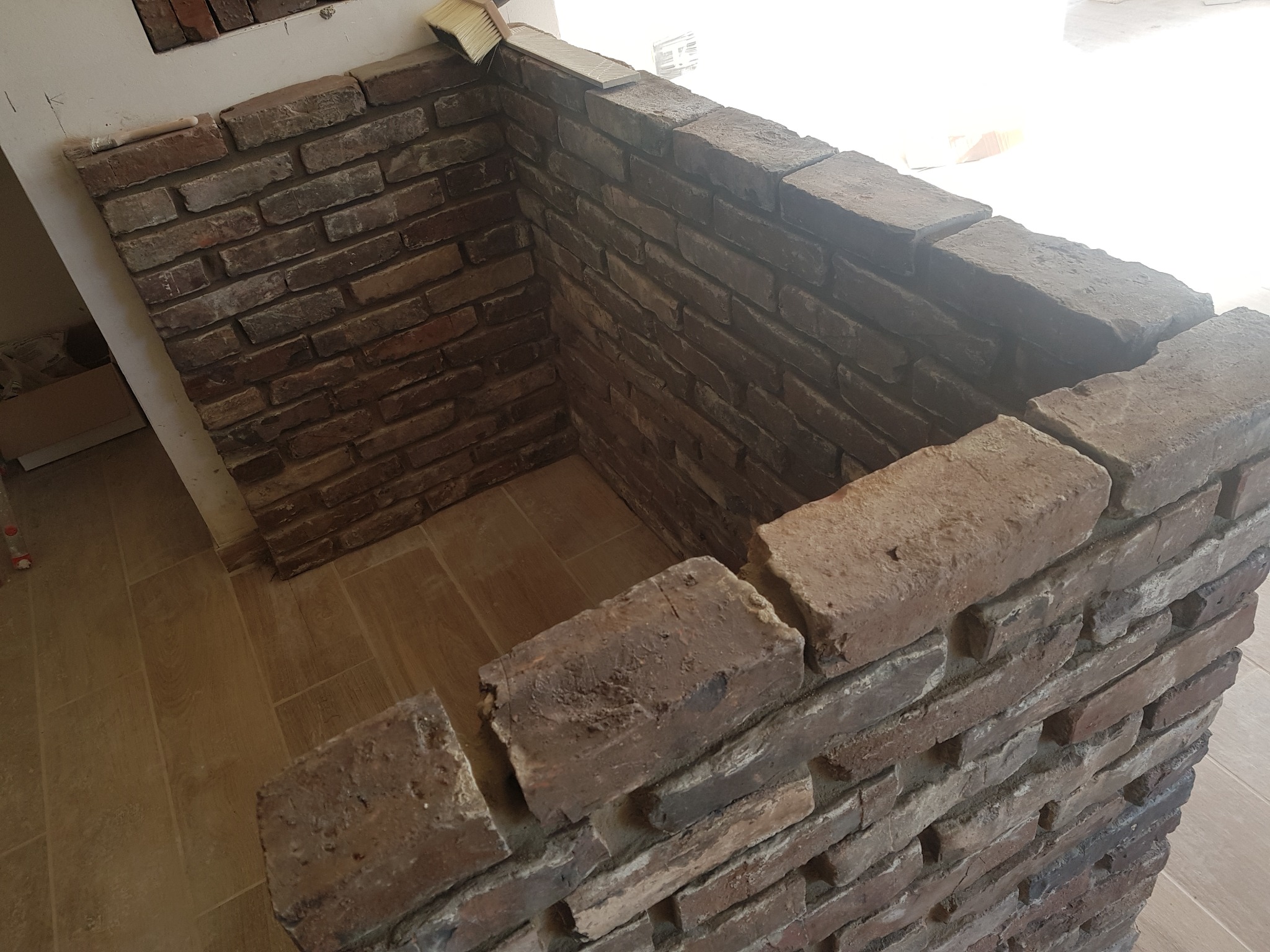 Fragment ściany wykończonej cegłą klinkierową w odcieniach brązu i szarości, z widoczną szczotką do fugowania na górnej krawędzi, na podłodze płytki imitujące drewno.