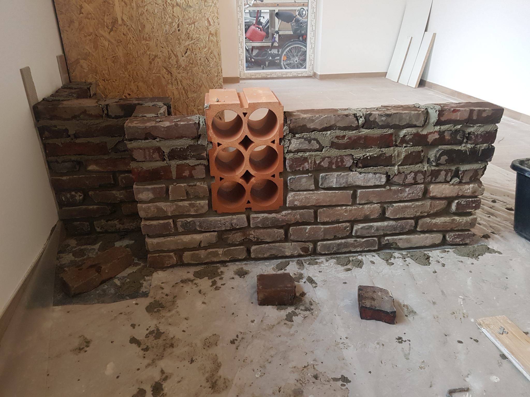 Murowana ścianka działowa z cegły klinkierowej w trakcie budowy, z wykorzystaniem pustaka ceramicznego z okrągłymi otworami, na surowym, zabrudzonym podłożu w jasnym pomieszczeniu.