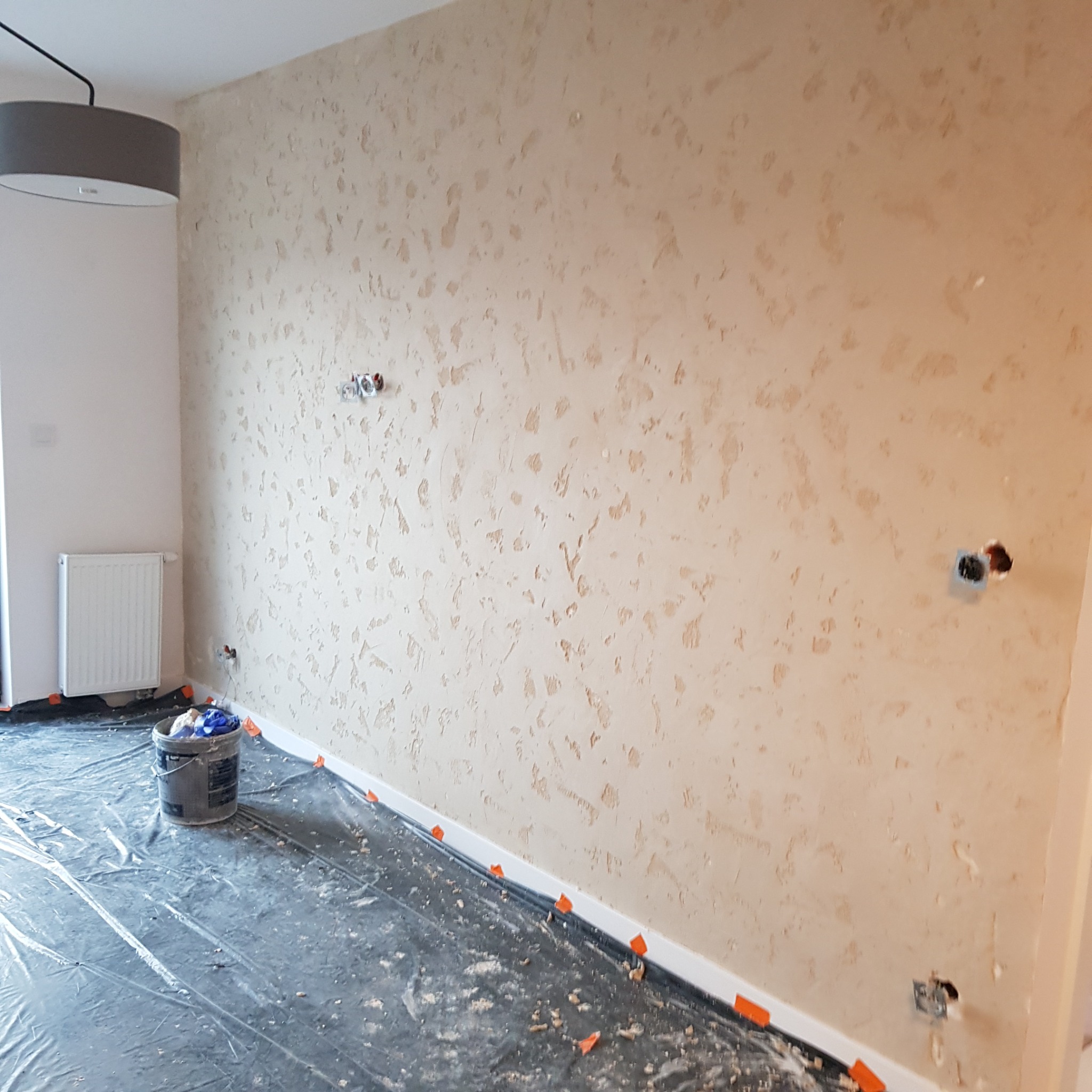 Pokój w trakcie nakładania dekoracyjnej struktury na ścianę, zabezpieczona podłoga folią, widoczny kubeł z materiałami i wystające puszki elektryczne.
