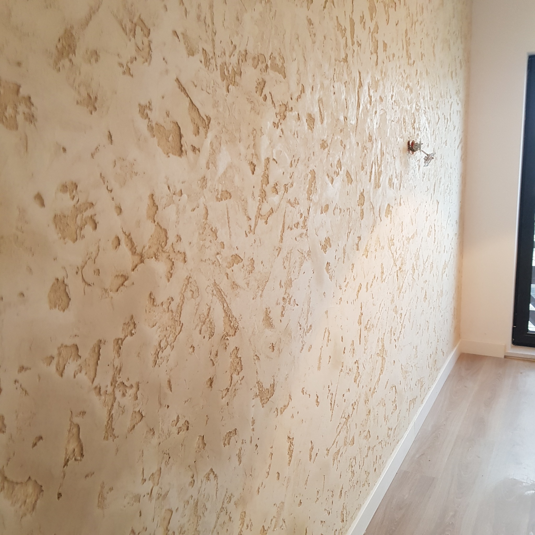 Ściana z dekoracyjną, beżową fakturą, imitującą naturalny kamień lub piaskowiec, z widocznymi gniazdkami elektrycznymi i fragmentem podłogi z jasnymi panelami.