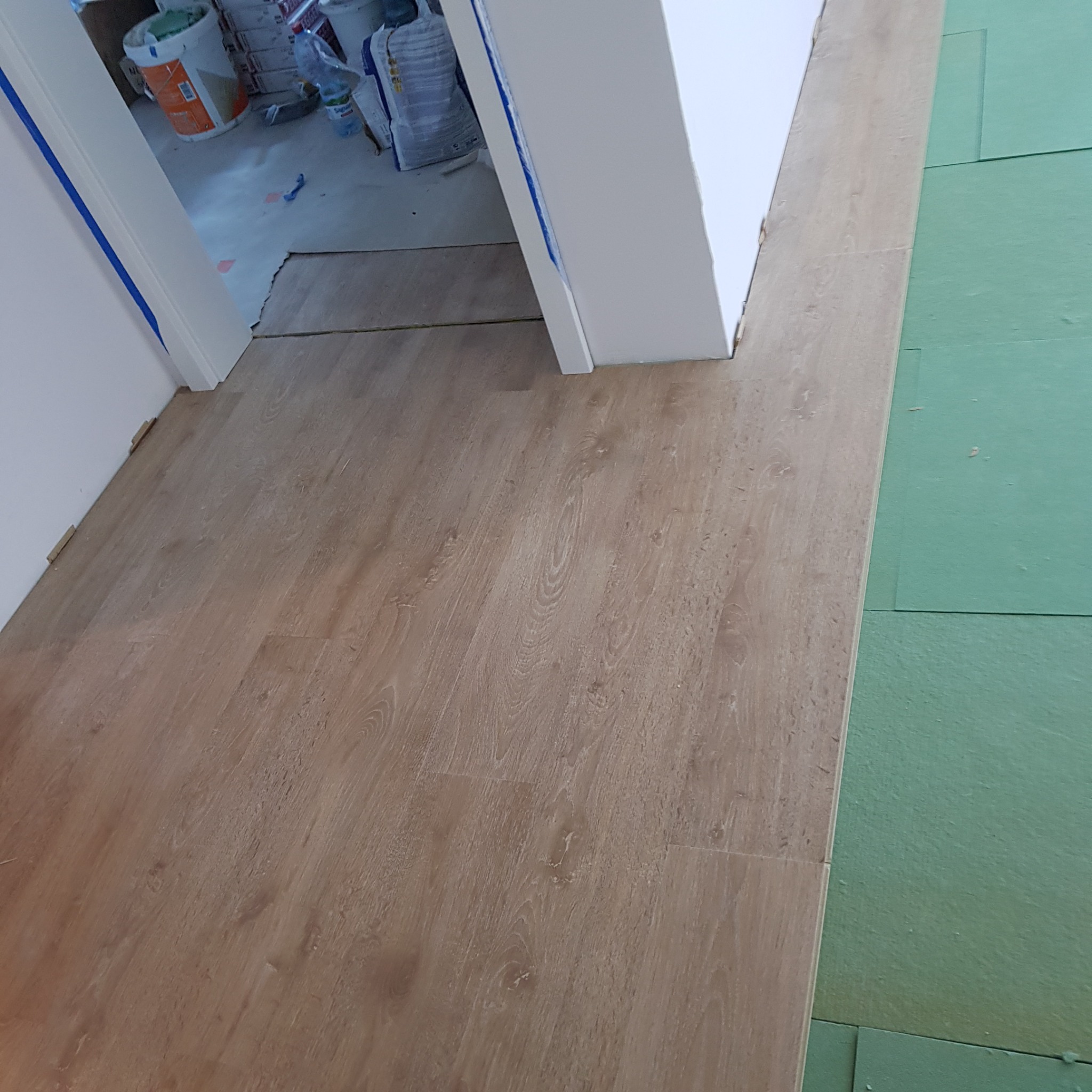 Fragment podłogi z paneli w odcieniach brązu, sąsiadujący z zieloną podkładką izolacyjną; widoczne również białe futryny drzwi zabezpieczone niebieską taśmą malarską.