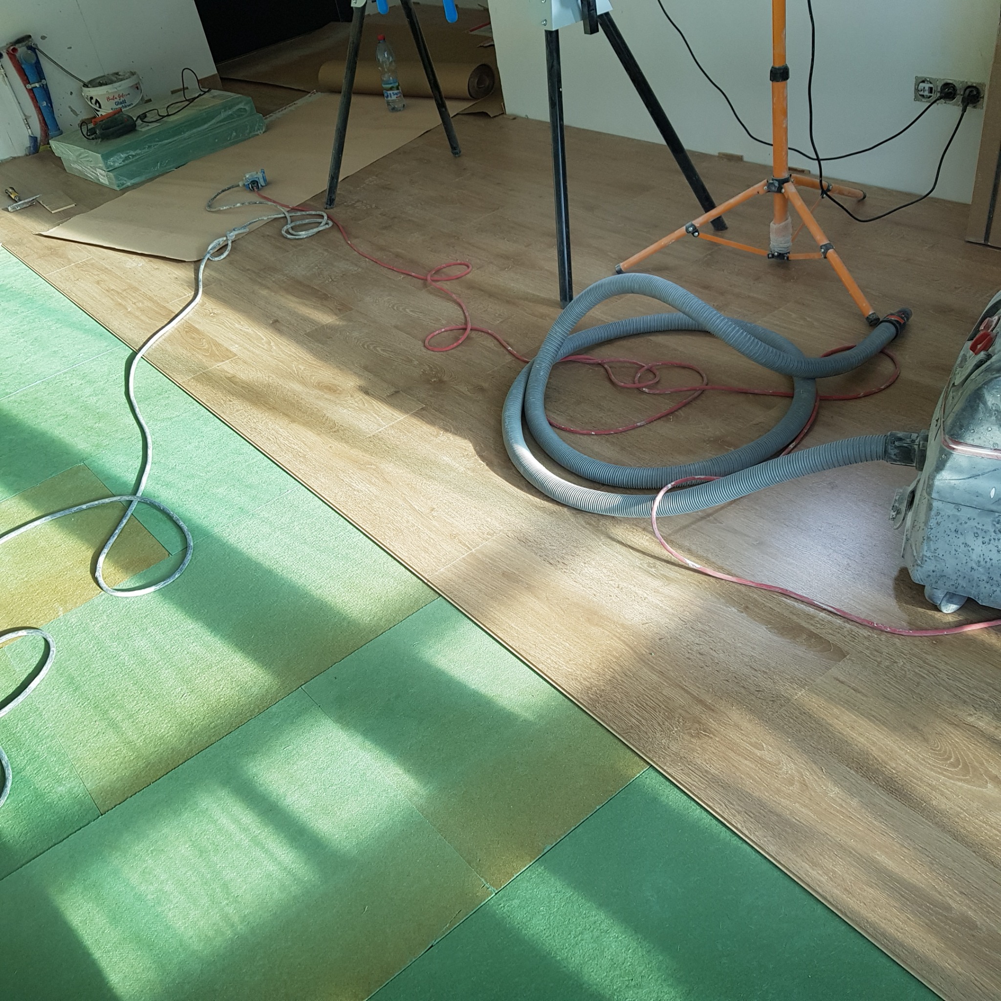 Fragment podłogi z ułożoną zieloną pianką podkładową i panelami w odcieniach brązu, widoczny odkurzacz przemysłowy z szarą rurą oraz lampa budowlana na trójnogu w tle.