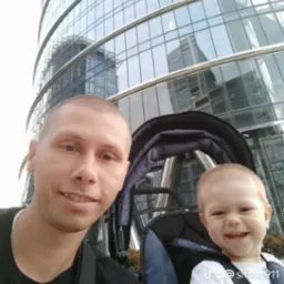 Uśmiechnięty mężczyzna z synem w w&oacute;zku na tle szklanego biurowca w słoneczny dzień.