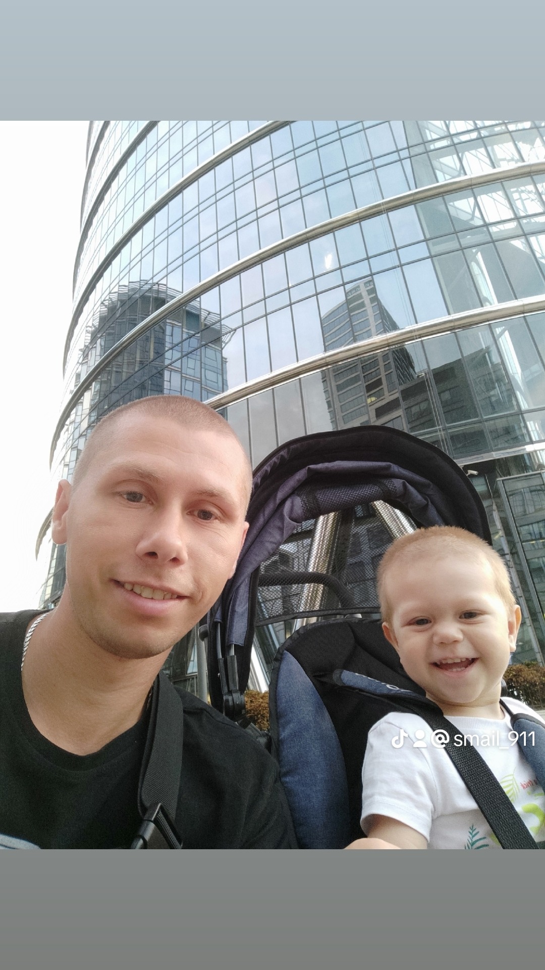 Uśmiechnięty mężczyzna z synem w wózku na tle szklanego biurowca w słoneczny dzień.