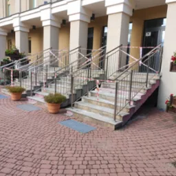 Schody wejściowe do budynku z metalowymi barierkami, zabezpieczone taśmą ostrzegawczą, z donicami z kwiatami po bokach, wyłożone czerwoną kostką brukową.