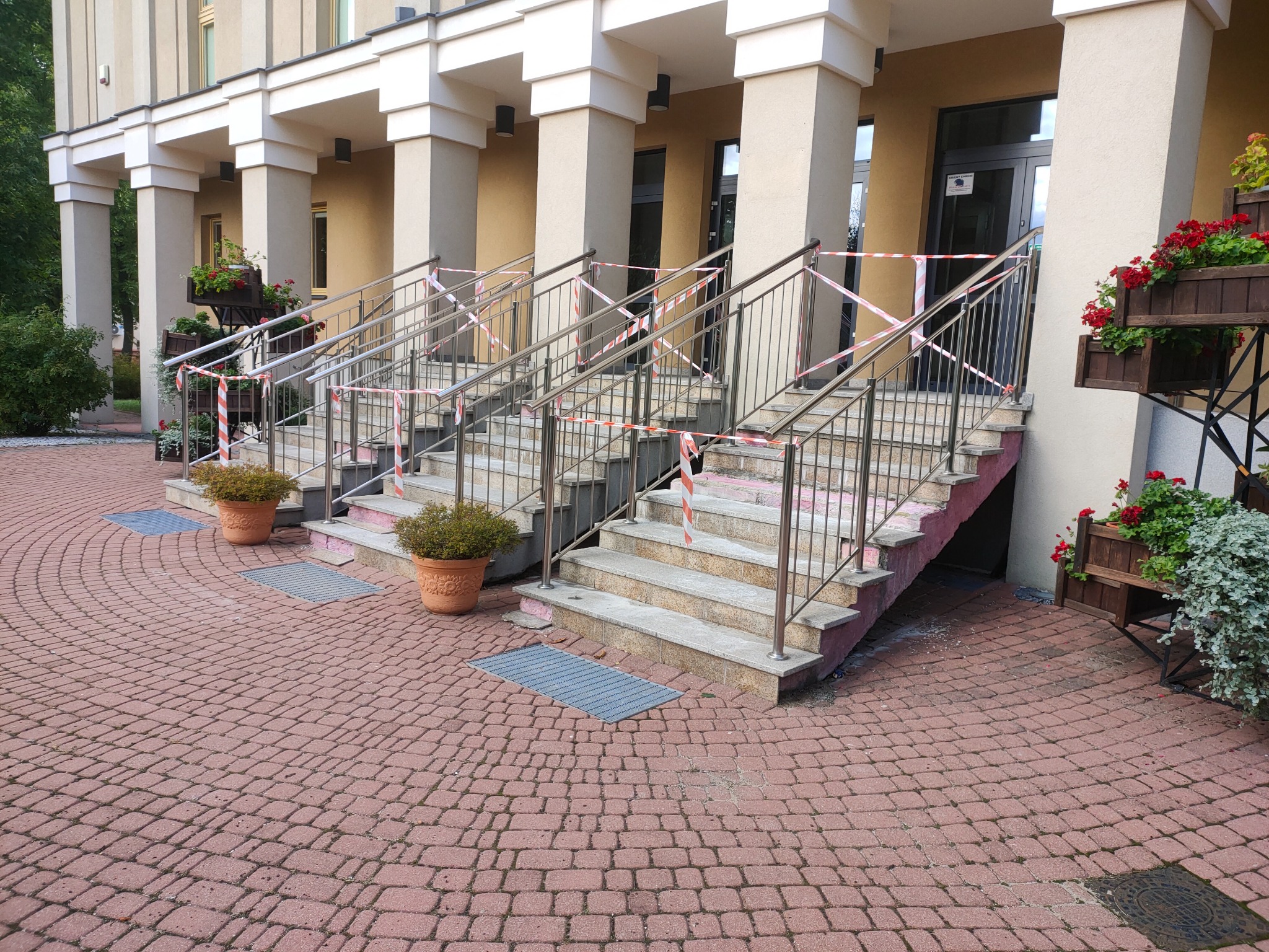 Schody wejściowe do budynku z metalowymi barierkami, zabezpieczone taśmą ostrzegawczą, z donicami z kwiatami po bokach, wyłożone czerwoną kostką brukową.