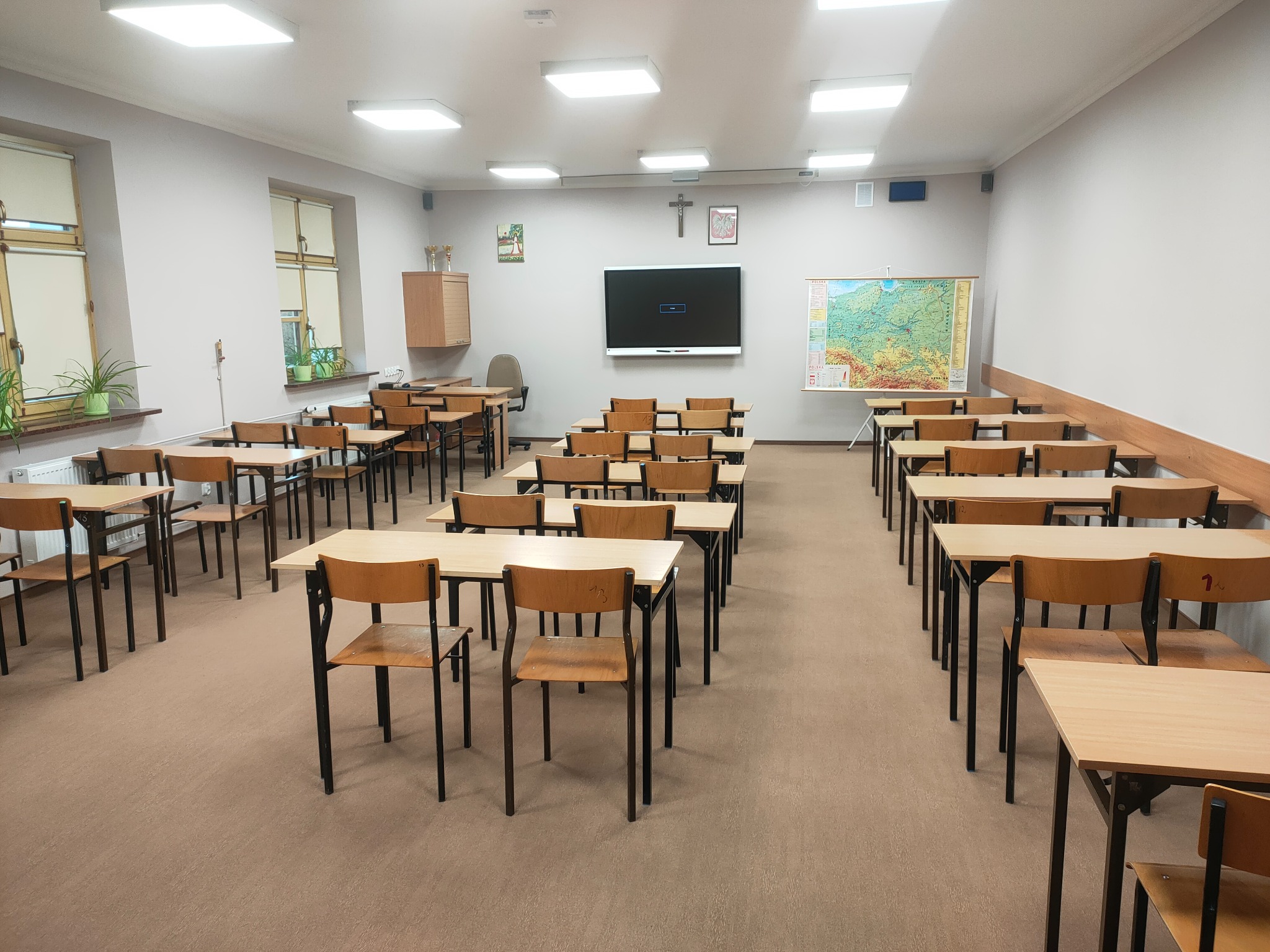 Jasna, przestronna sala lekcyjna z rzędami drewnianych ławek i krzeseł, tablicą interaktywną, mapą Polski na ścianie i oświetleniem sufitowym.