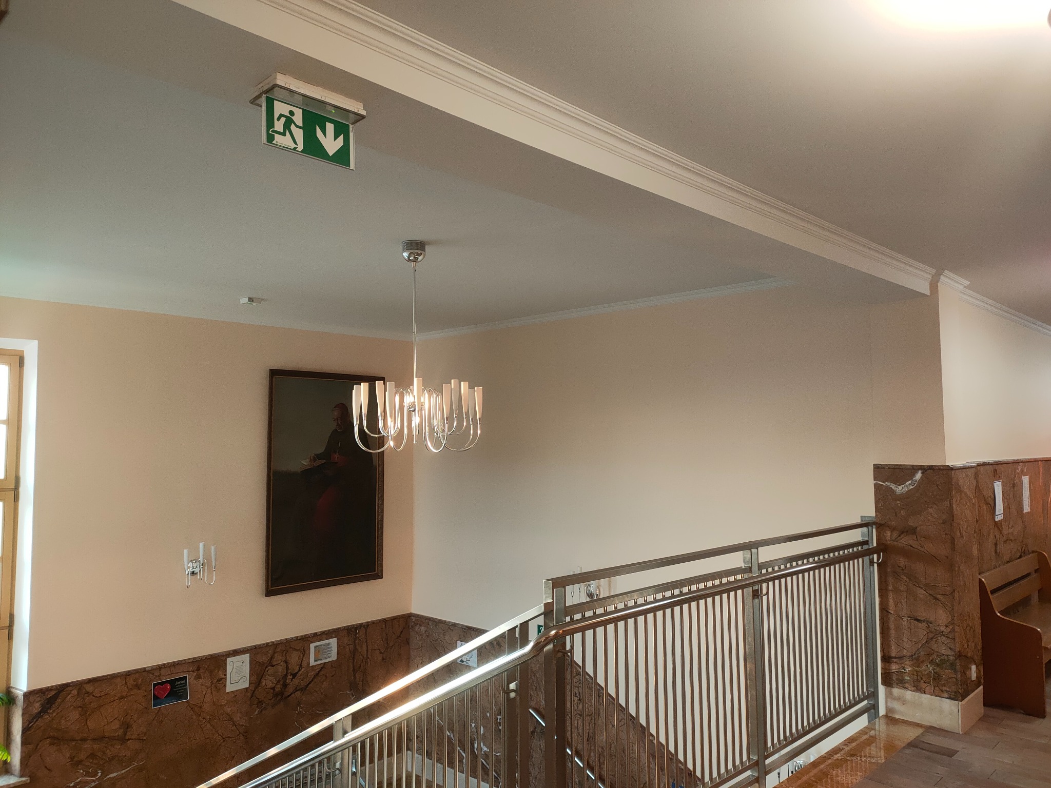 Klatka schodowa z marmurowymi ścianami, obrazem w złotej ramie, nowoczesnym żyrandolem i znakiem ewakuacyjnym na suficie.