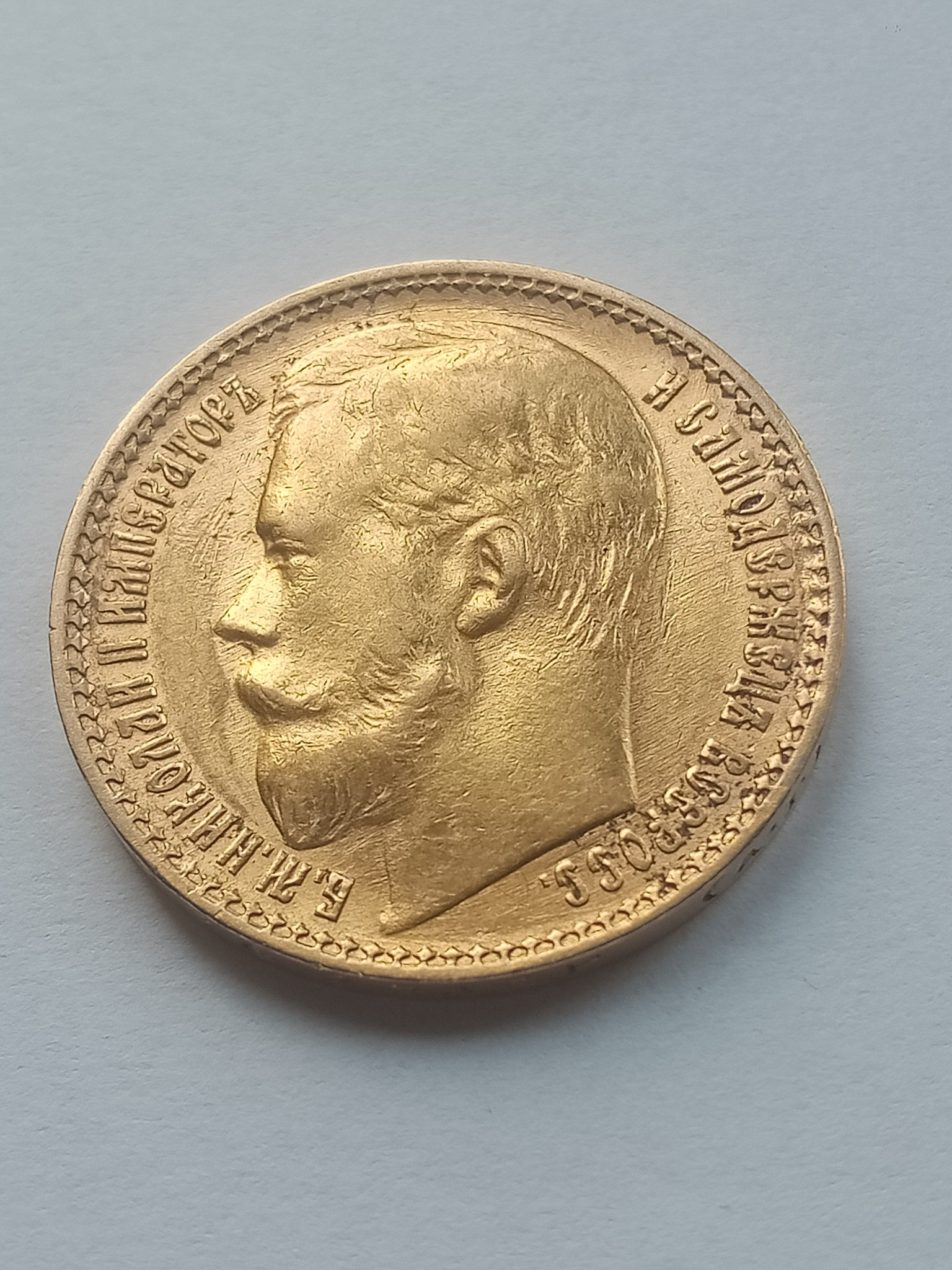 Złota moneta z wizerunkiem cara Mikołaja II na jasnym tle, widoczny profil z wyraźnym wąsem i inskrypcje w języku rosyjskim wokół krawędzi.