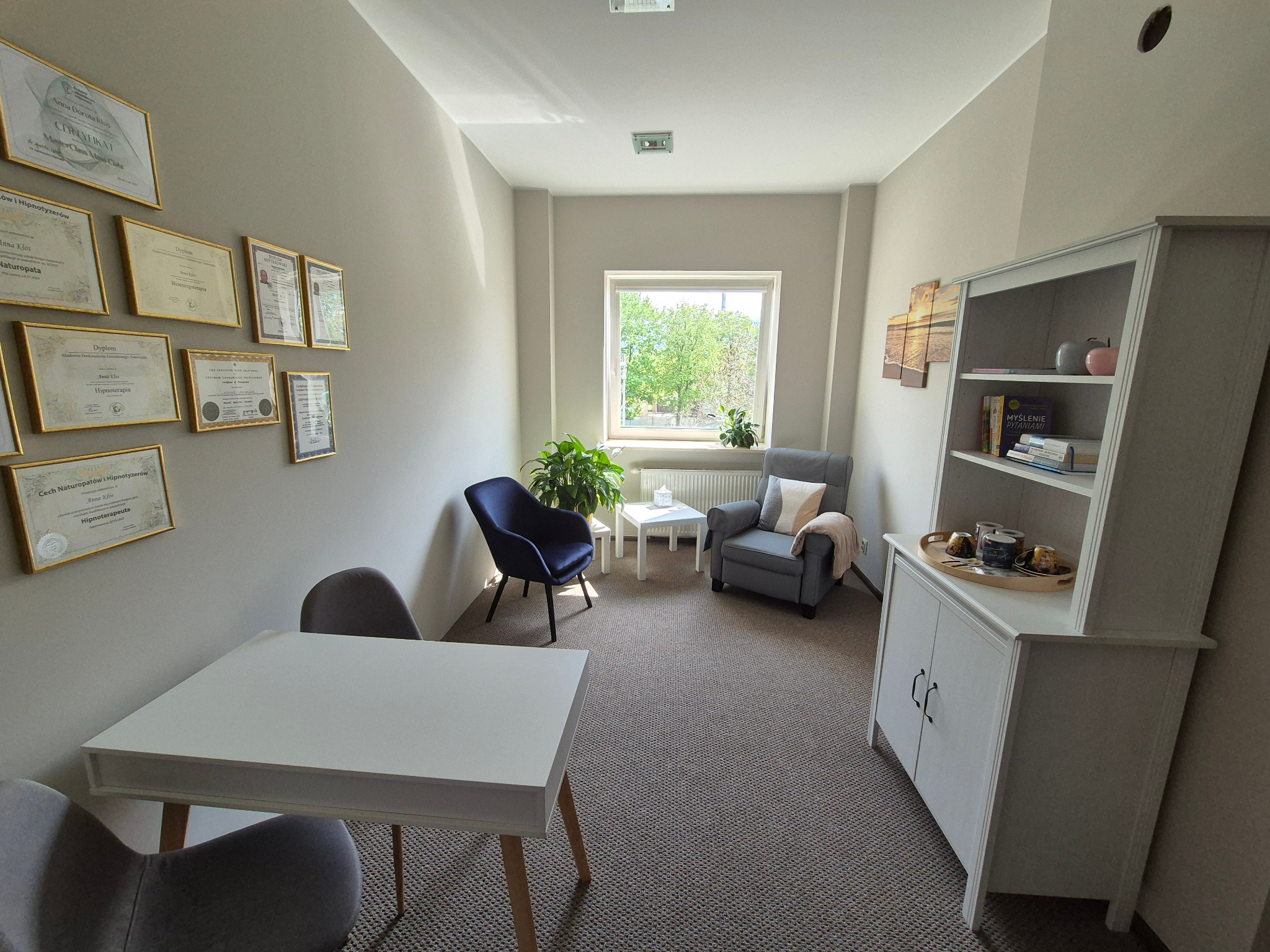 Jasne wnętrze gabinetu terapeutycznego z dyplomami na ścianie, białym biurkiem i dwoma krzesłami, w głębi fotel i stolik kawowy przy oknie z zielenią.