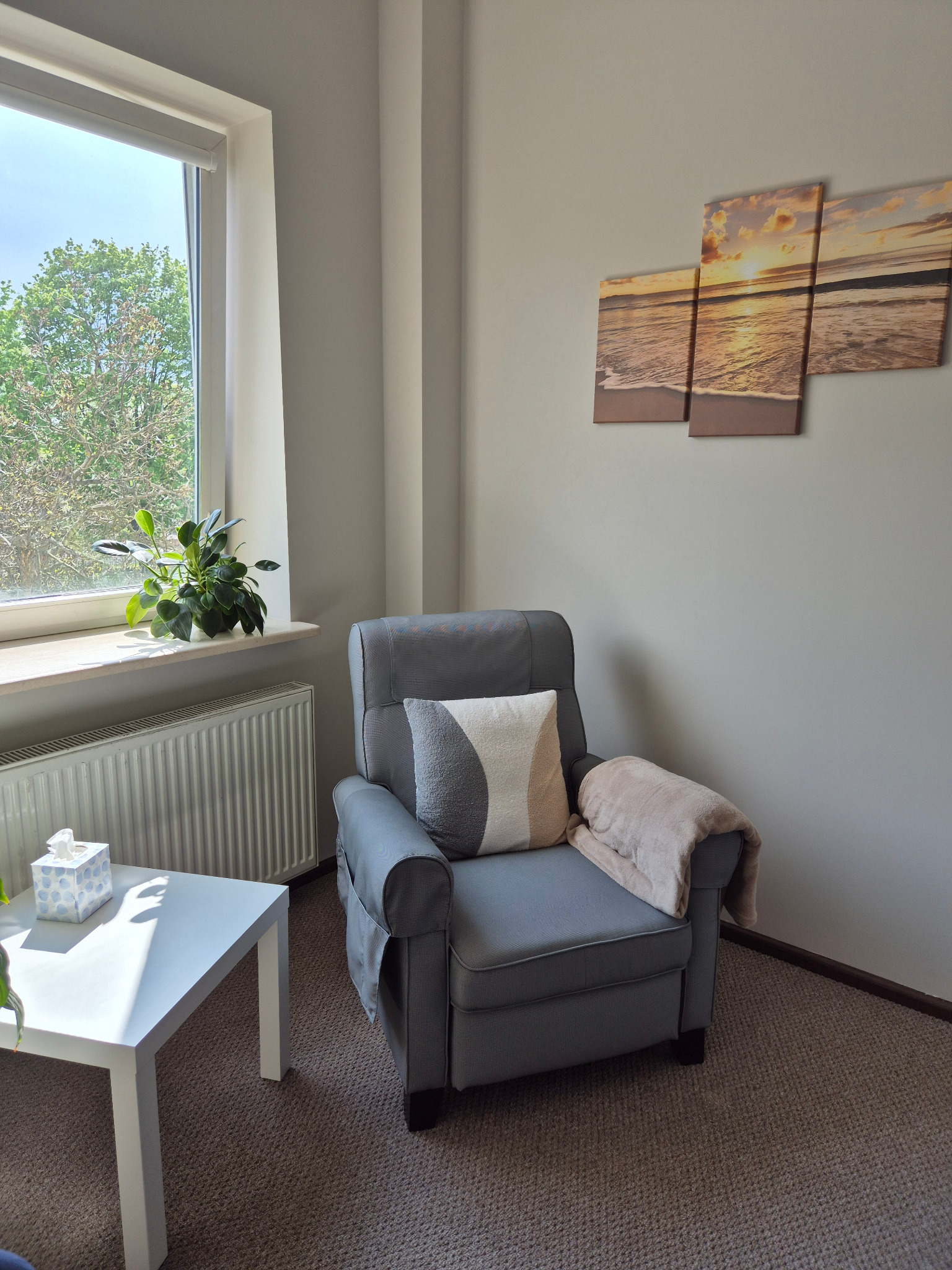 Jasne, minimalistyczne wnętrze gabinetu z szarym fotelem, beżowym pledem, dekoracyjną poduszką, stolikiem z chusteczkami, kwiatem w doniczce na parapecie i obrazem morza nad fotelem.