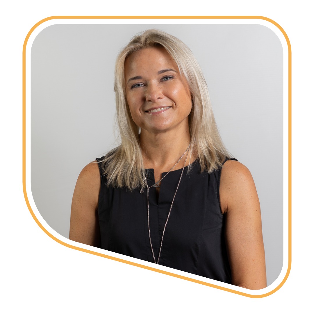 Portret uśmiechniętej blondynki w czarnej sukience z dwoma srebrnymi łańcuszkami na jasnym tle.