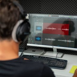WoogieStudio49 - Osoba w słuchawkach edytuje ścieżki audio na ekranie komputera w wygłuszonym pomieszczeniu, widoczne fale dźwiękowe i mikser w programie do obróbki audio.