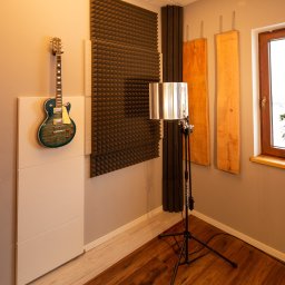 WoogieStudio49 - Wnętrze studia nagrań z gitarą elektryczną wiszącą na ścianie, panele akustyczne piramidkowe i drewniane, mikrofon na statywie z filtrem pop, drewniana podłoga, roślina w doniczce na parapecie.