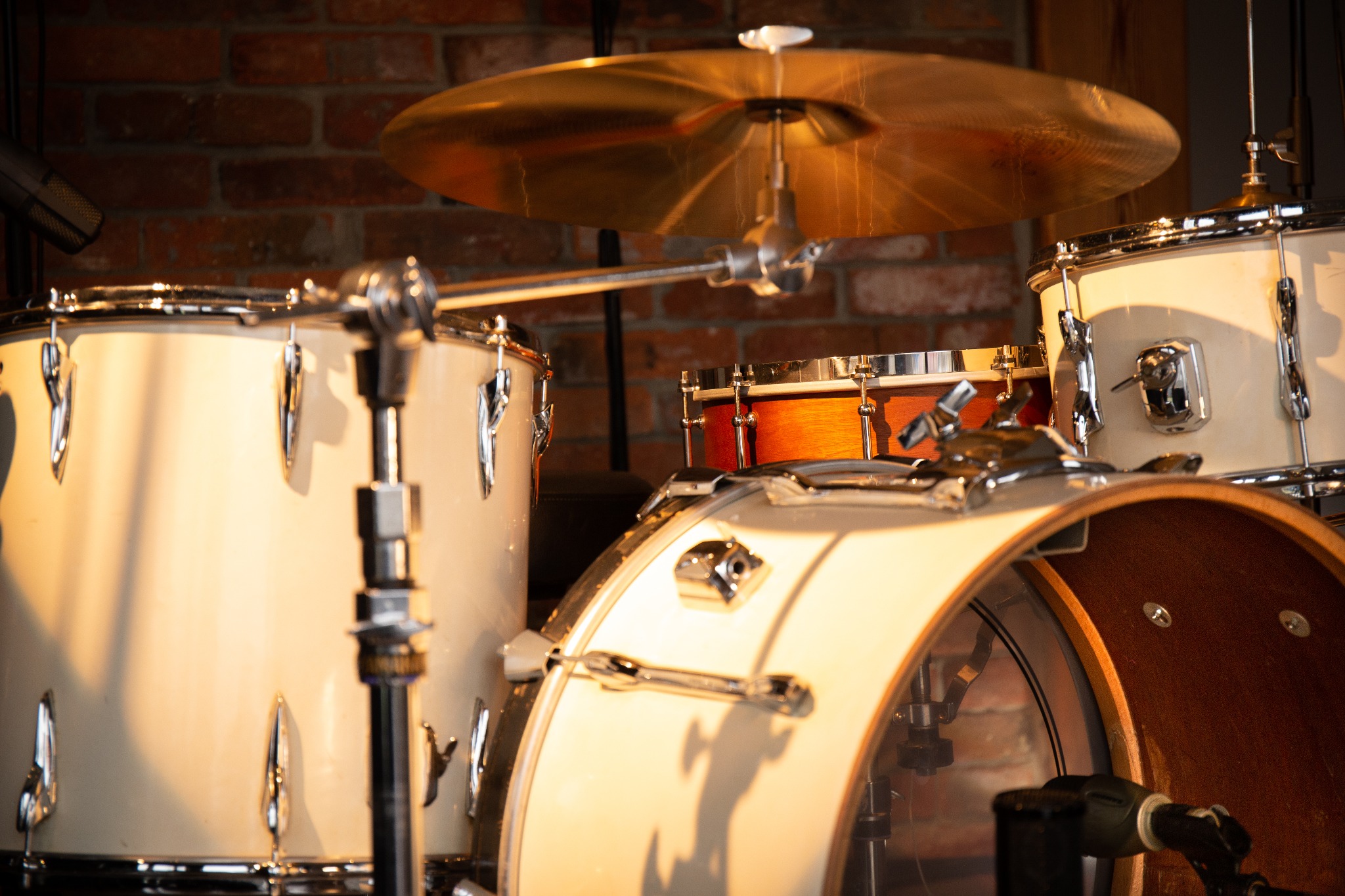 Zestaw perkusyjny z białymi bębnami i mosiężnym talerzem na tle ściany z czerwonej cegły, oświetlony ciepłym światłem.