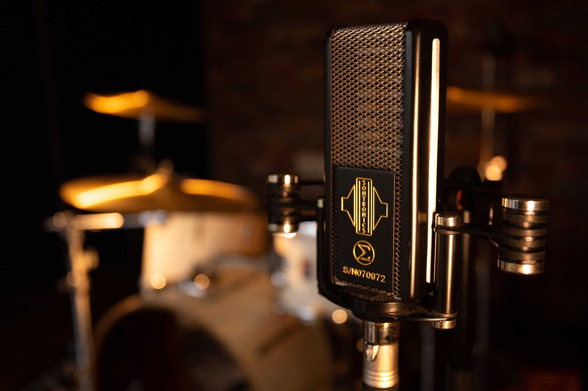 Czarny mikrofon pojemnościowy Sontronics z metalową siatką ochronną i logo na tle rozmytej perkusji