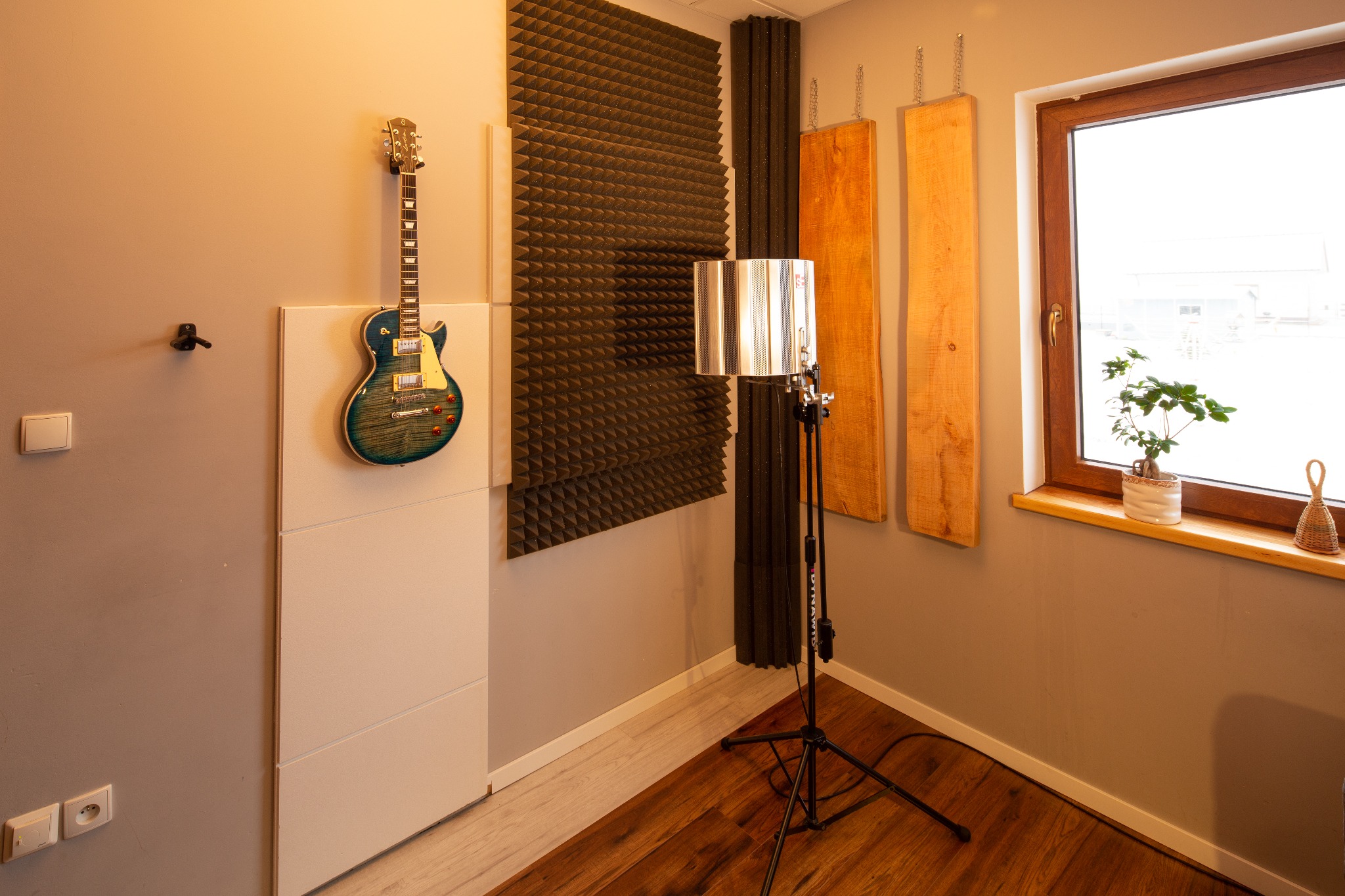 Wnętrze studia nagrań z gitarą elektryczną wiszącą na ścianie, panele akustyczne piramidkowe i drewniane, mikrofon na statywie z filtrem pop, drewniana podłoga, roślina w doniczce na parapecie.