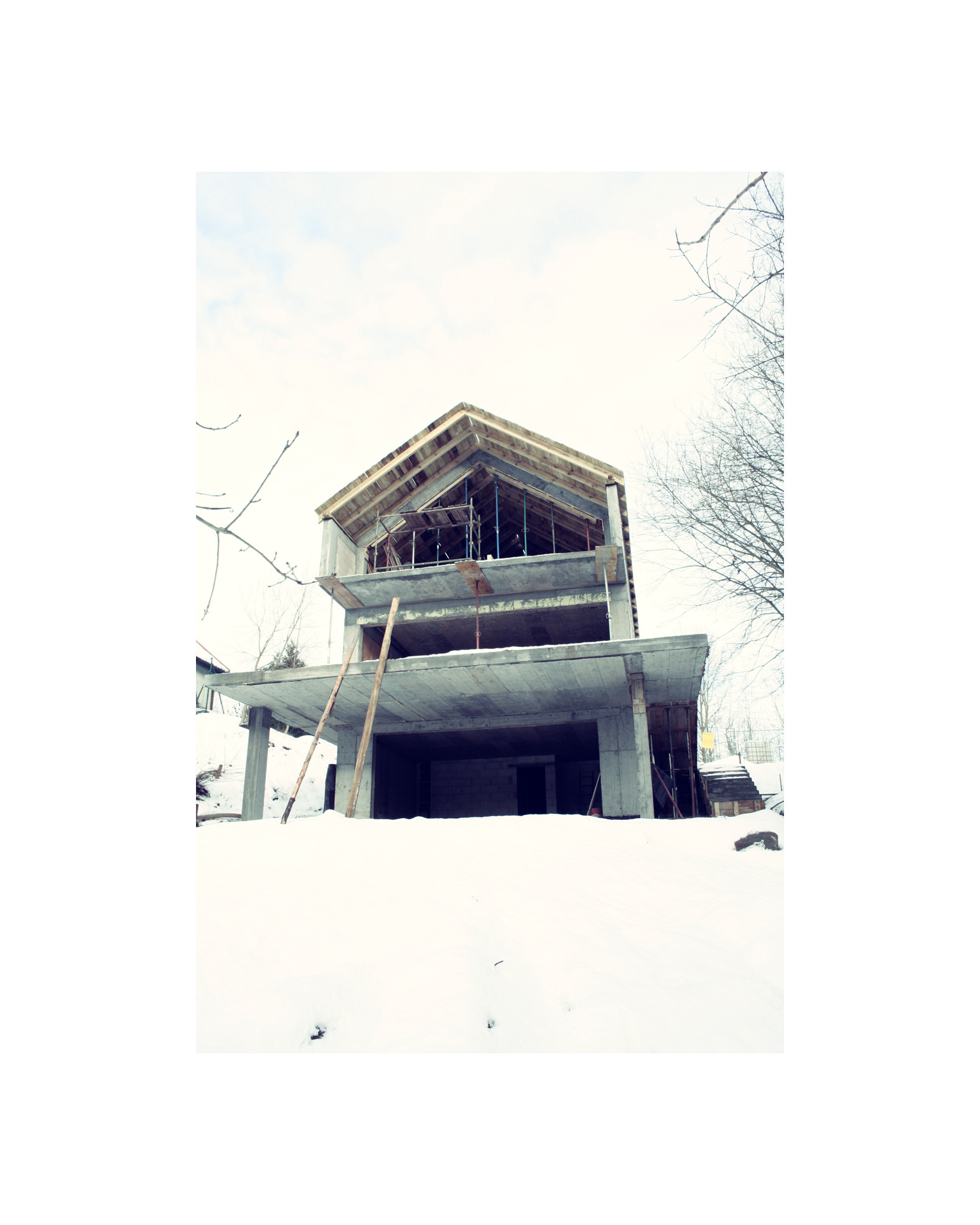 Surowy stan domu w trakcie budowy, widok z dołu w zimowej scenerii, z drewnianą konstrukcją dachu i betonowymi elementami.