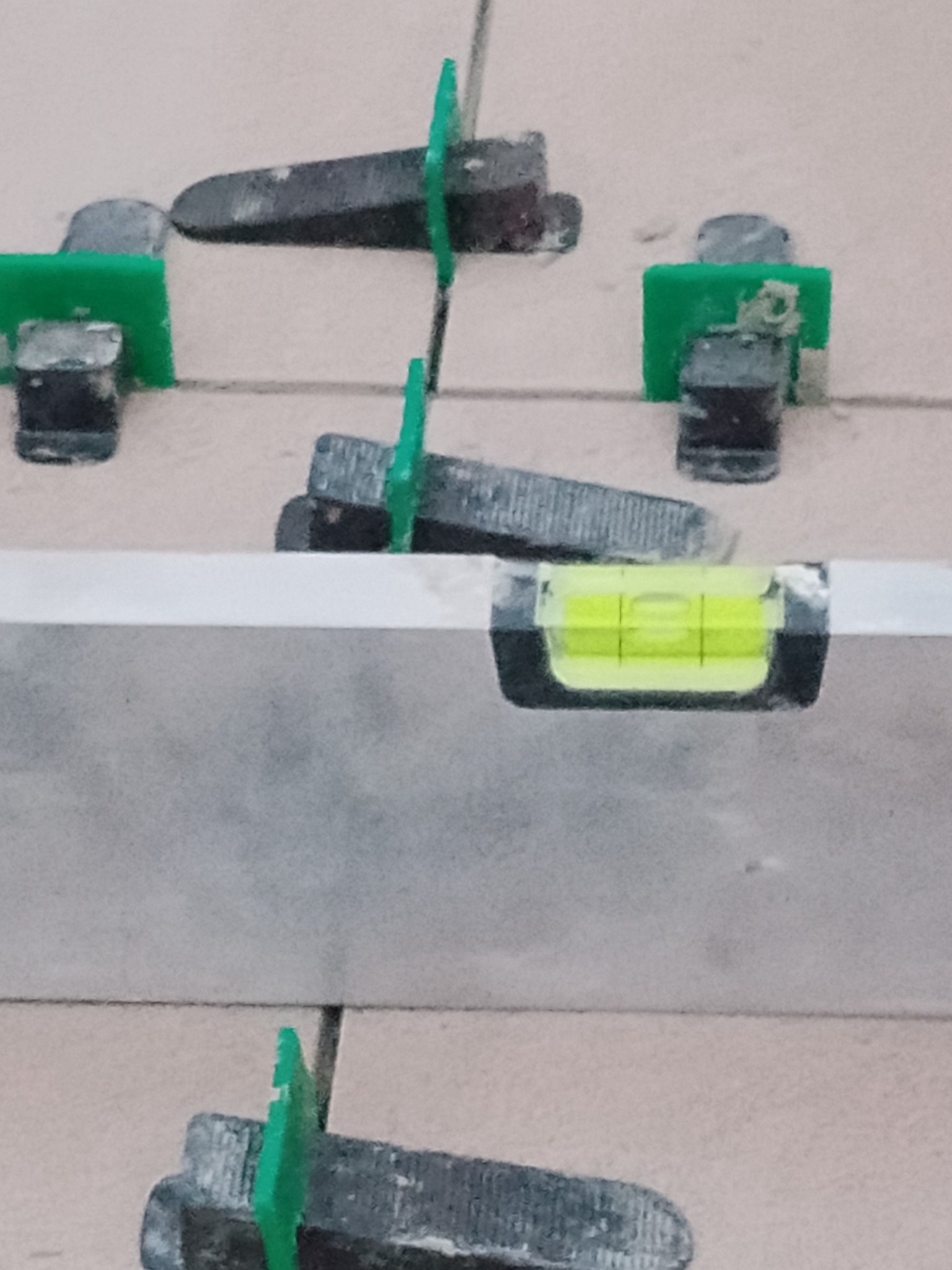 Poziomica i system poziomowania płytek z zielonymi klinami i czarnymi klipsami na jasnych płytkach. Detal z bliska, ukazujący precyzję układania.