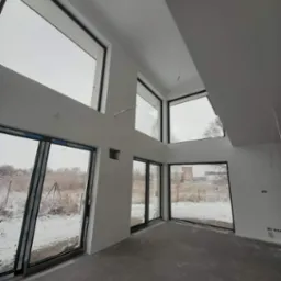 Wnętrze domu w stanie deweloperskim z dużymi oknami z czarnymi ramami, widok na zaśnieżony krajobraz, surowy beton na podłodze, białe ściany przygotowane do dalszych prac.